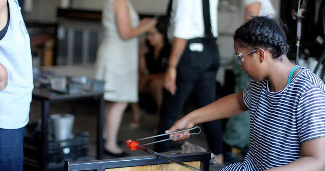 A young girl wearing safety glasses and a striped shirt is using tongs to hold a flaming object over heat indoors while other people are blurred in the background.