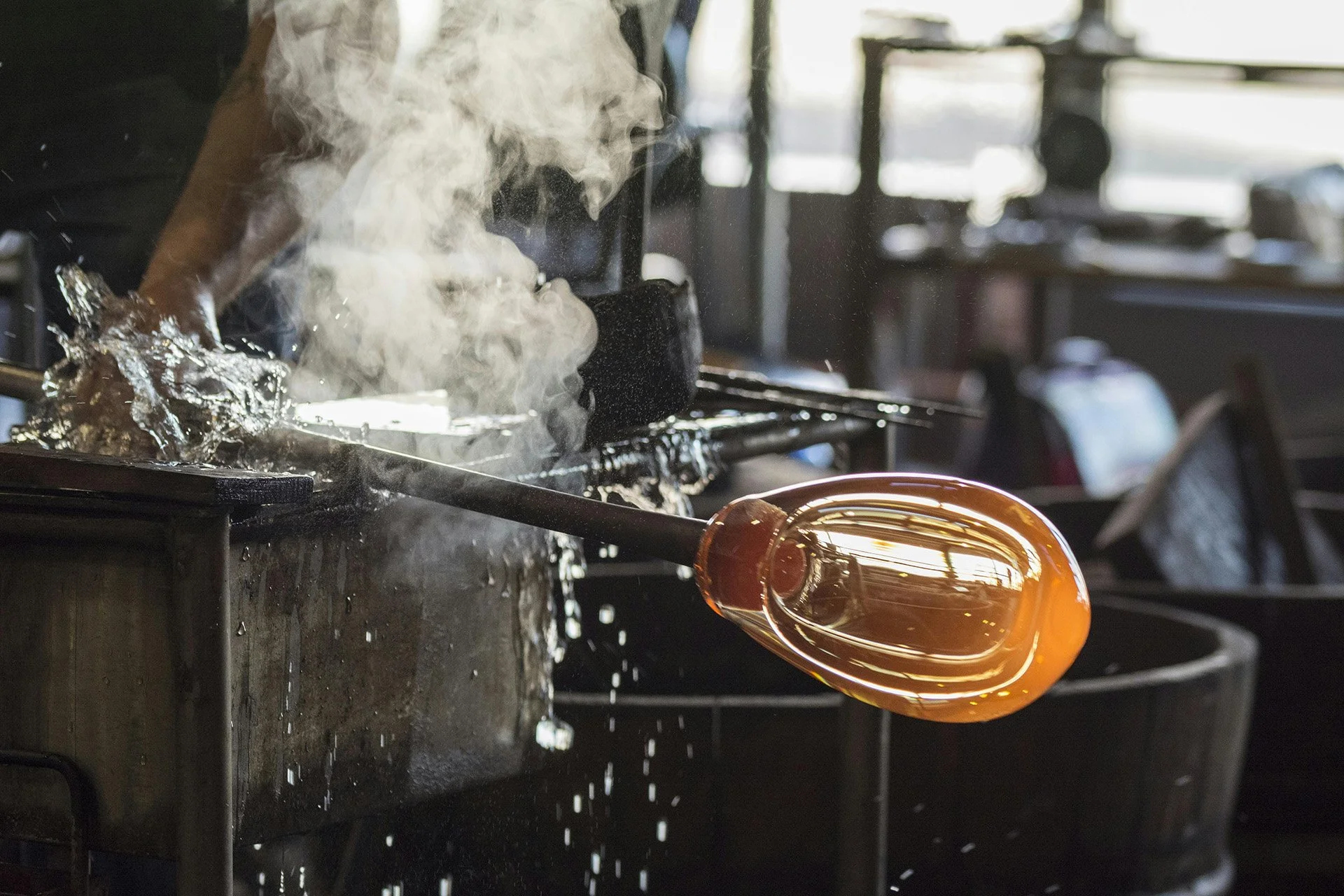 A glassblowing artist shaping molten glass in a workshop with steam and tools visible.