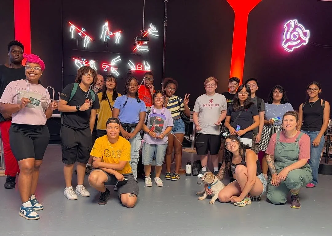 A group of diverse young people posing together indoors, some holding small items, with neon and LED lights on a dark wall behind them.