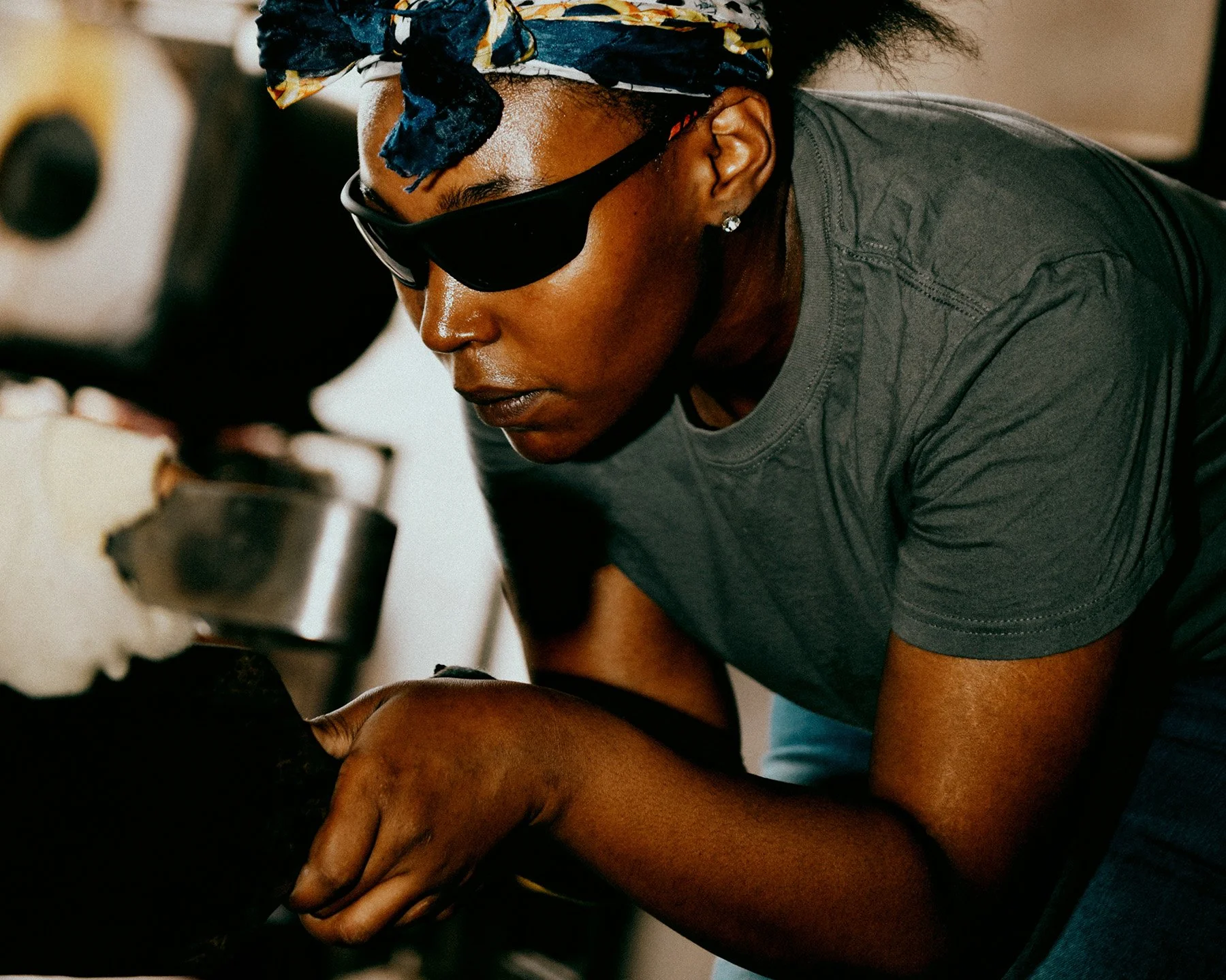 A woman wearing sunglasses and a bandana, focused on repairing a car engine.