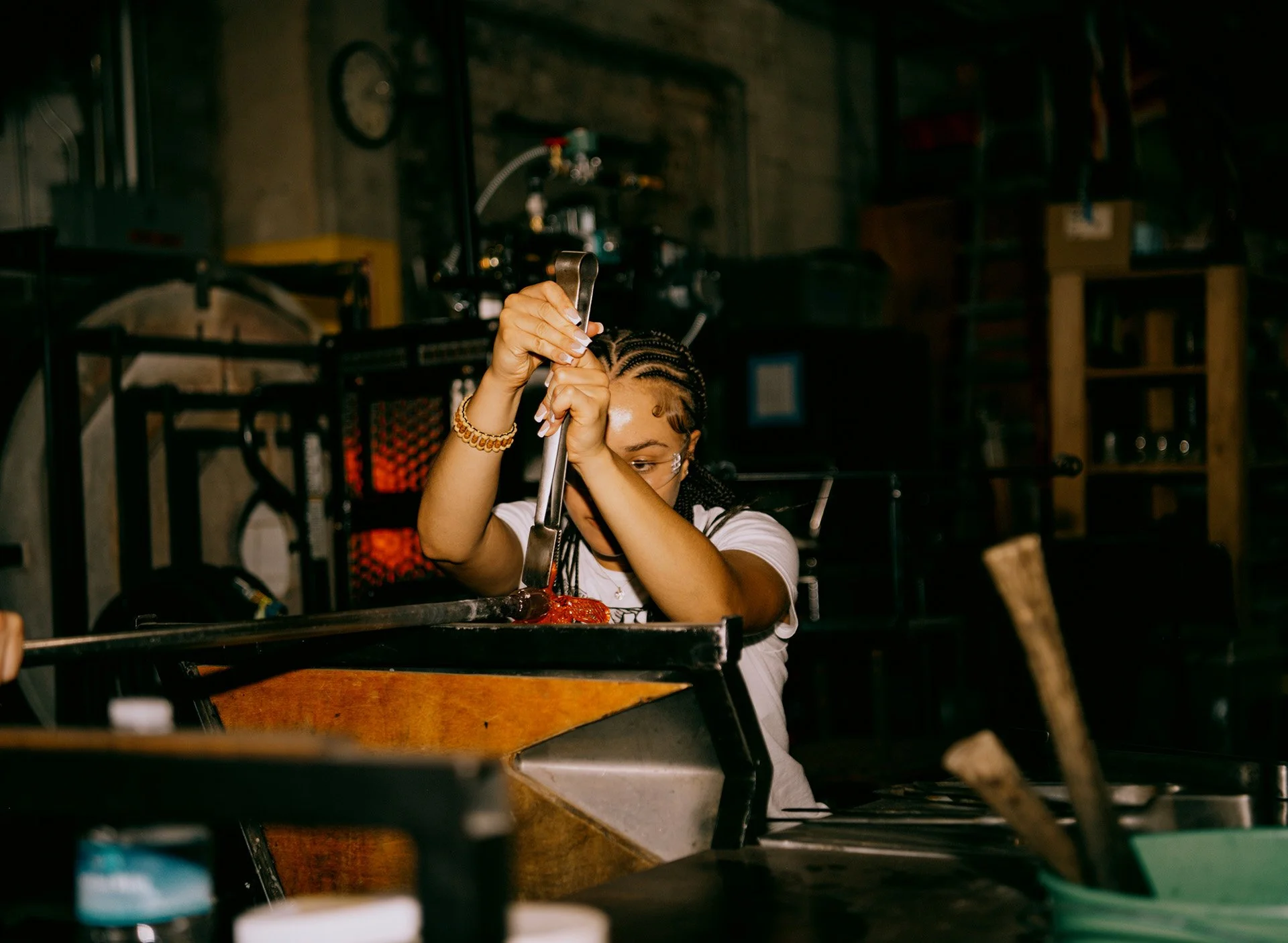 A person with braided hair wearing a white shirt and bracelets, working with tongs on a piece of meat in a kitchen or workshop, with various tools and equipment around.