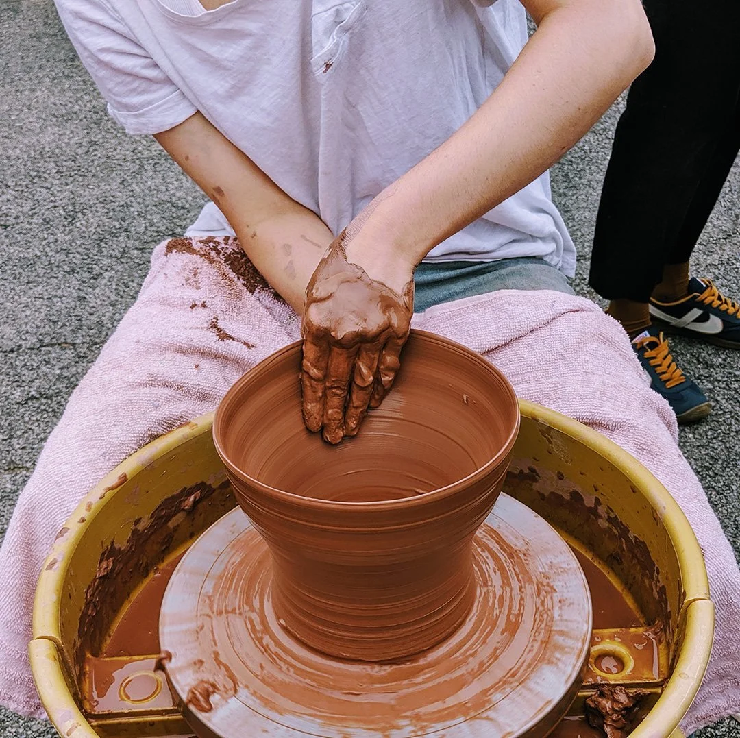 Person shaping a clay pottery bowl on a spinning wheel with muddy hands, nearby on a pink cloth.