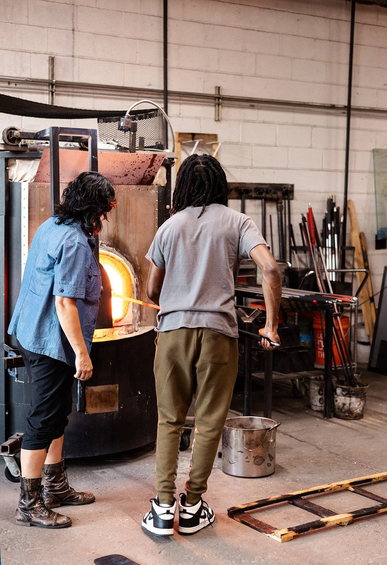 Two people working in a glassblowing studio, using a furnace to shape molten glass.