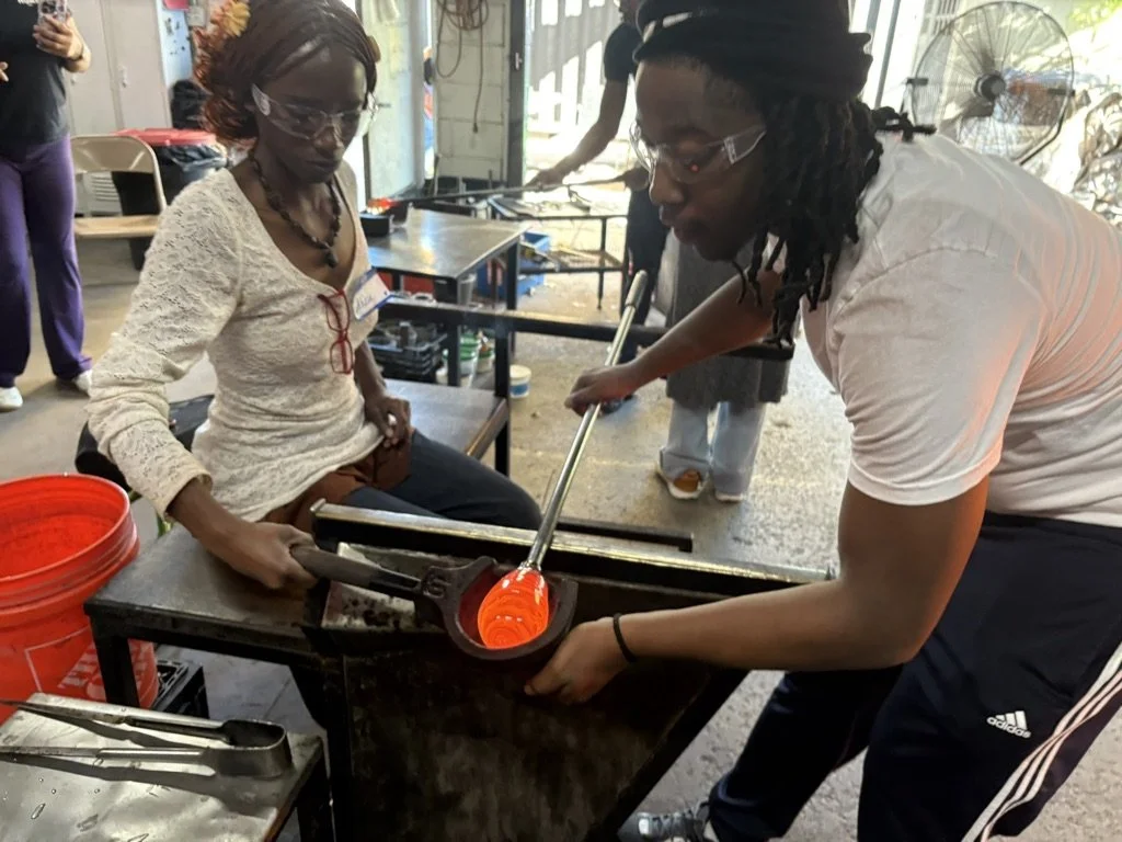 A glassblower is shaping molten glass using a blowpipe while a woman observes closely. They are working in a workshop with tools and safety equipment.