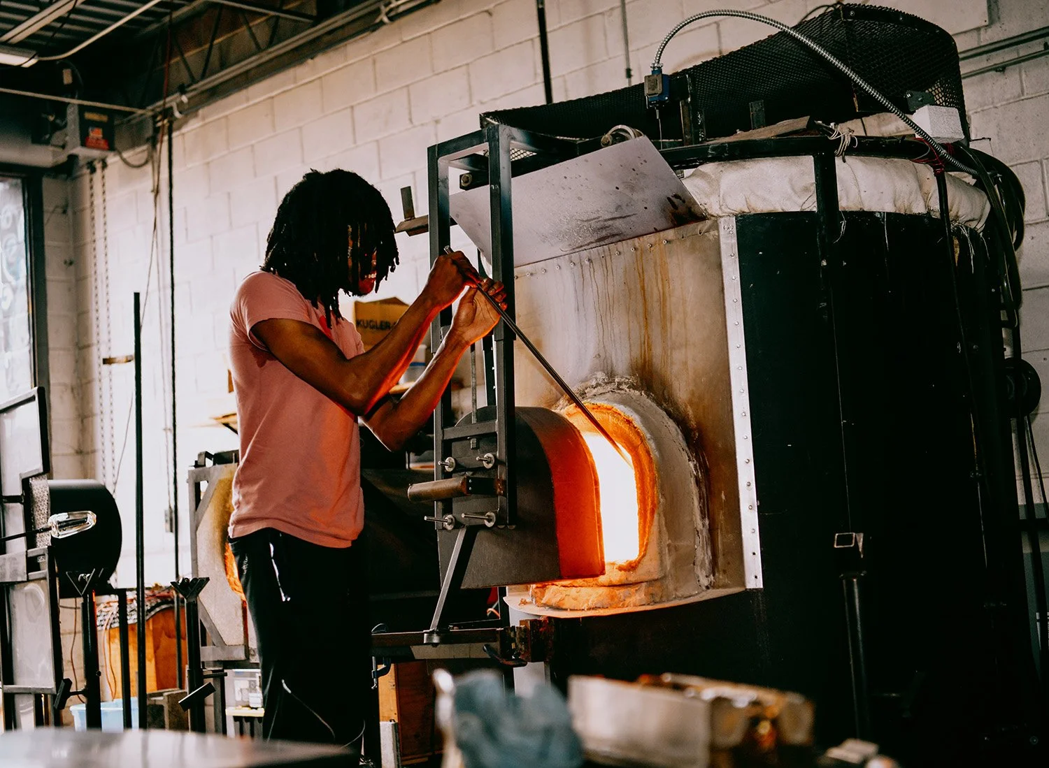A person with dreadlocks working with a glassblowing furnace in an industrial workshop.