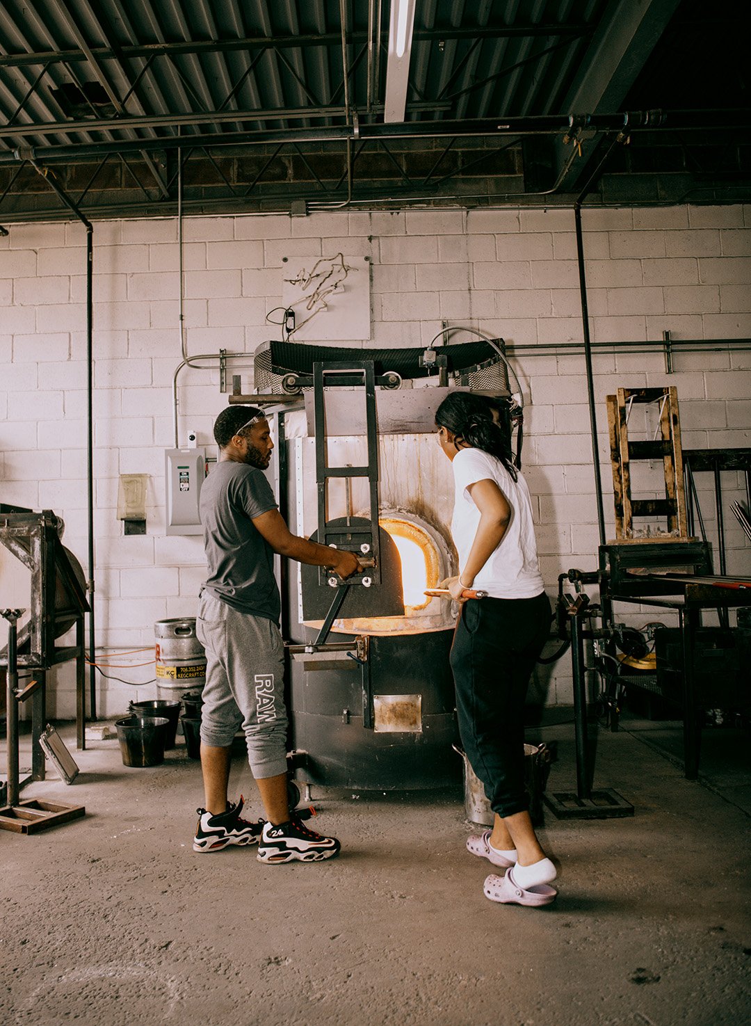 Two people stand at a furnace for glassblowing, blowing into a pipe to form glasswork.
