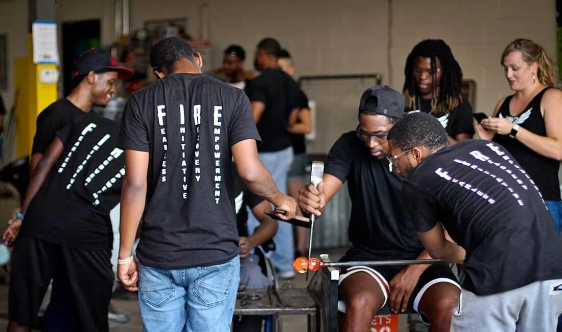 Group of young people in black shirts at a glassblowing workshop, some working with glass objects and others observing, in an industrial studio setting.