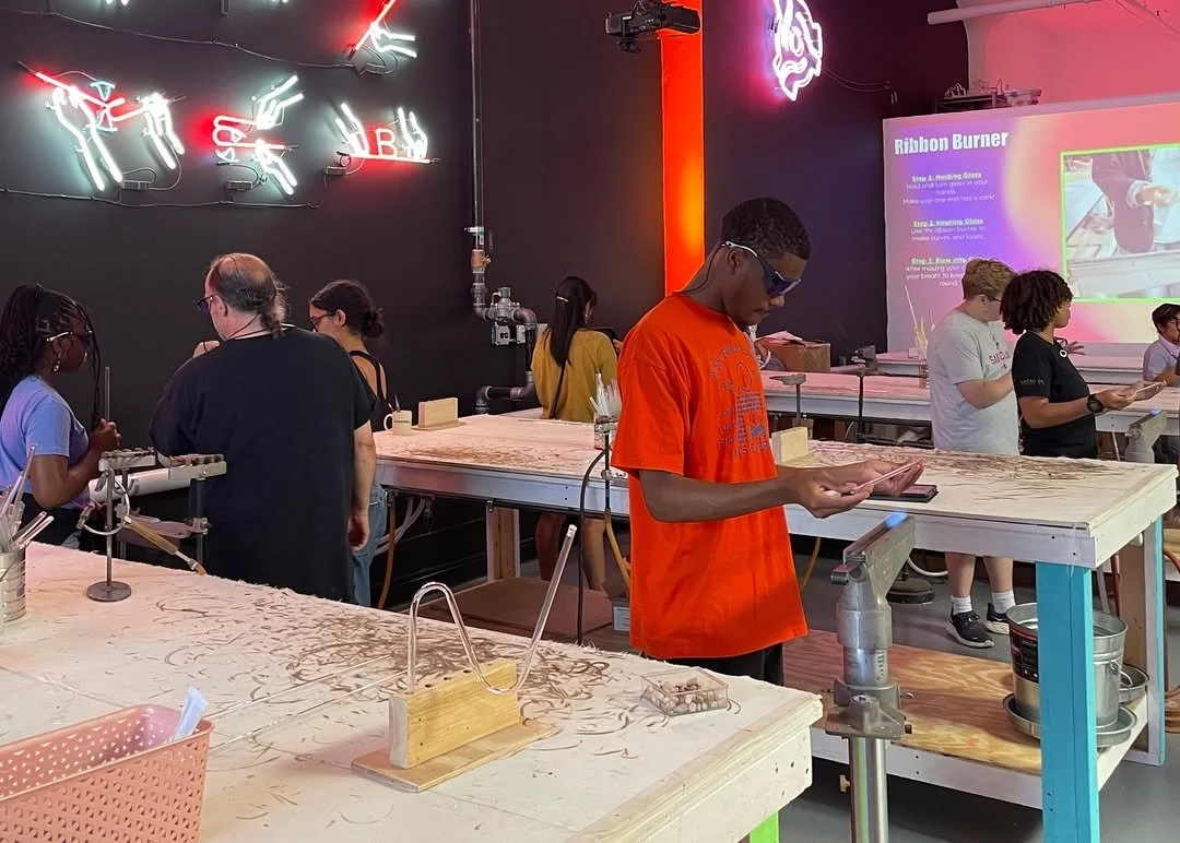 A group of people are participating in a woodworking or craft workshop inside a room with black walls decorated with neon signs and a neon horse head. They are working at long work tables with tools and materials, some using saws and carving tools. 