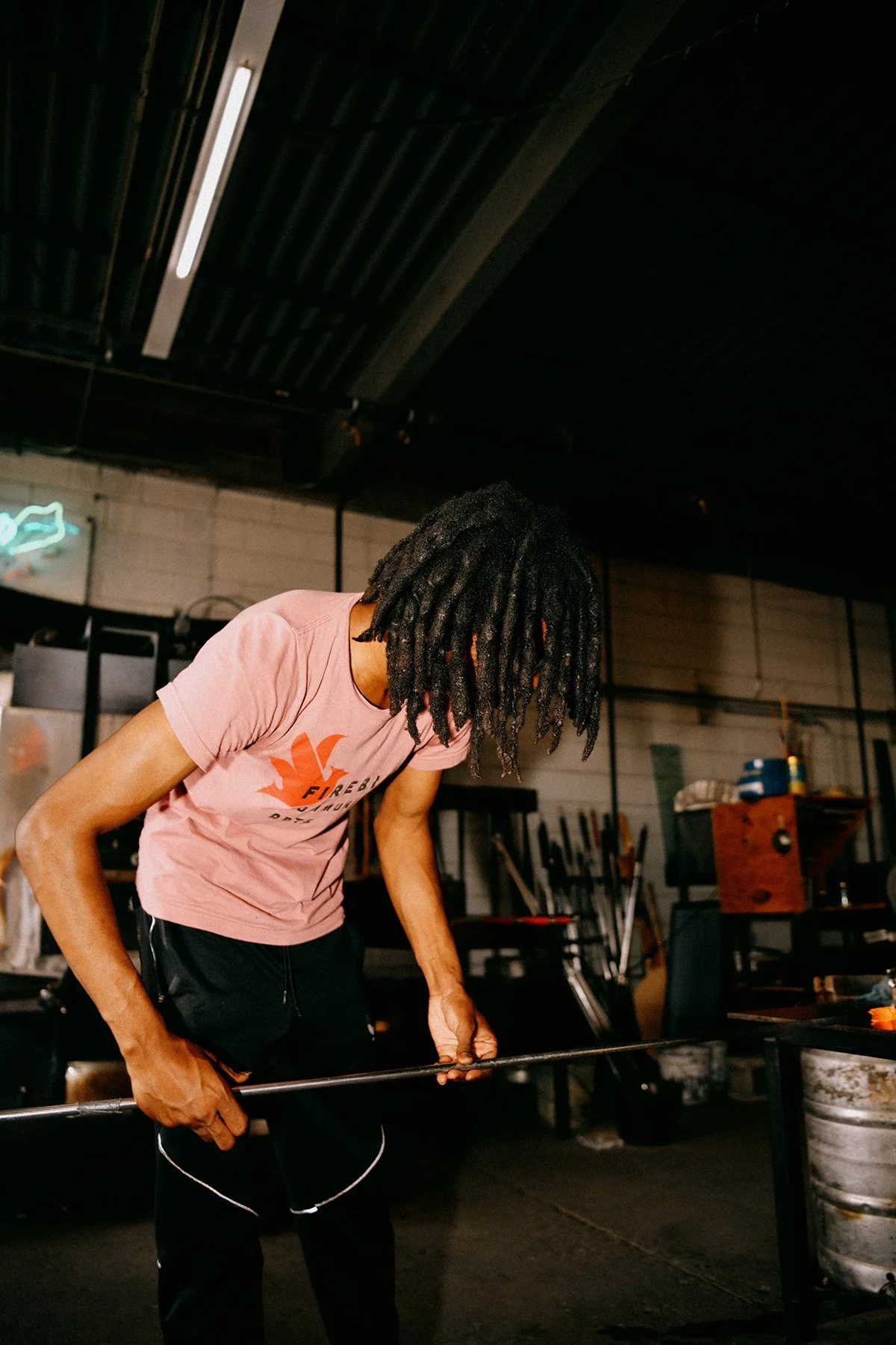 A man with dreadlocks wearing a pink T-shirt and black shorts is working in a workshop, holding a metal rod and leaning over a table.