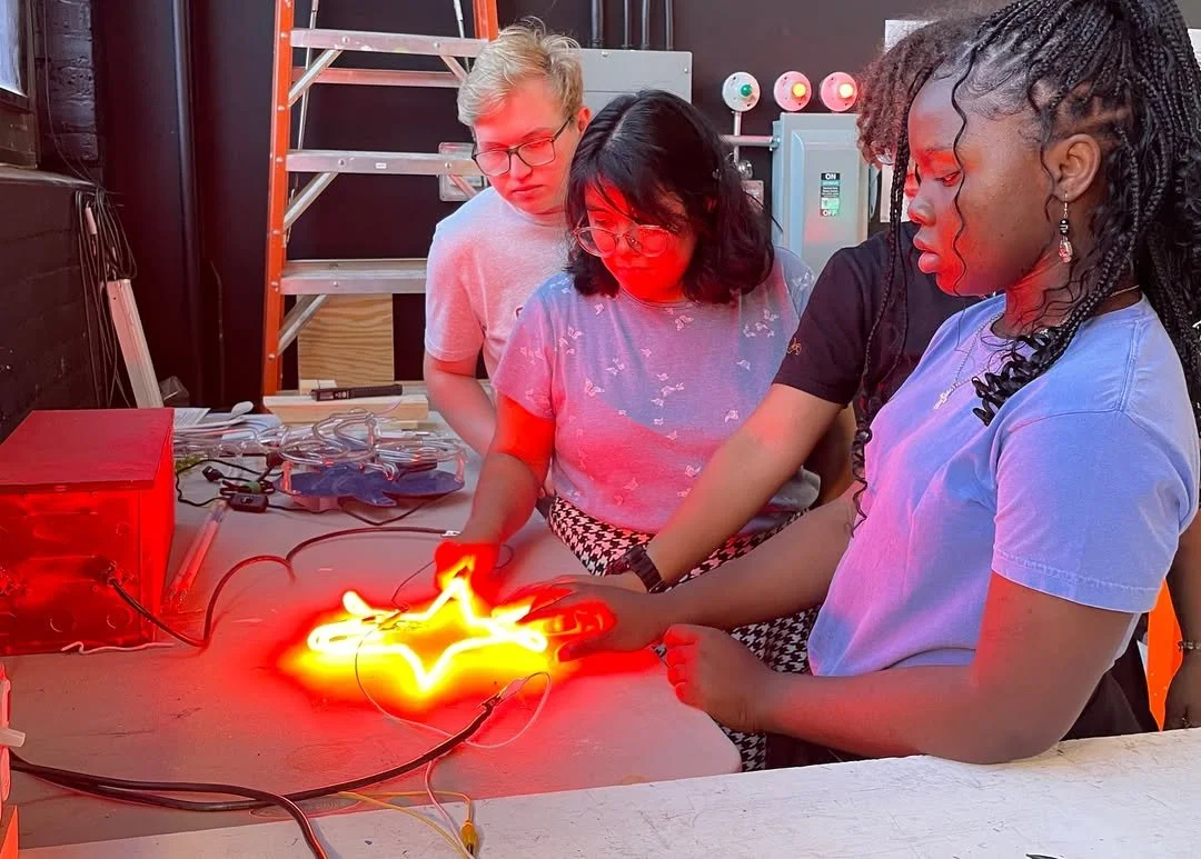 Group of four students working with a glowing neon star sign and electronic equipment on a table in a workshop.