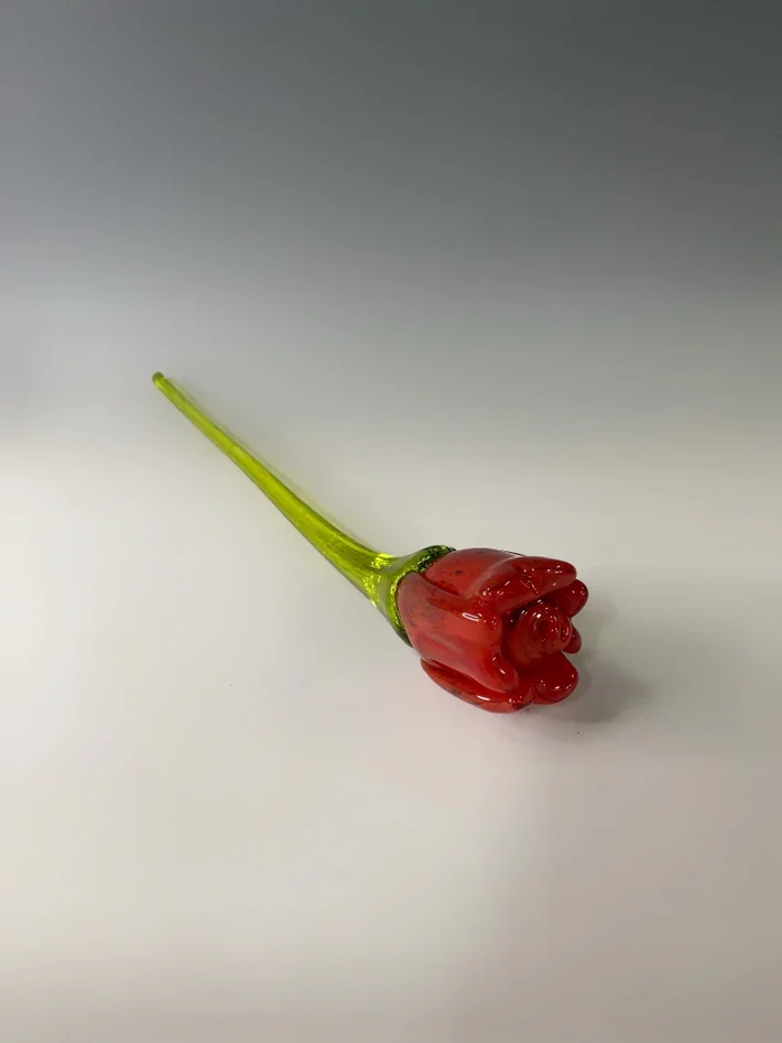 A small decorative glass flower with a red bloom and a green stem, lying on a white surface.