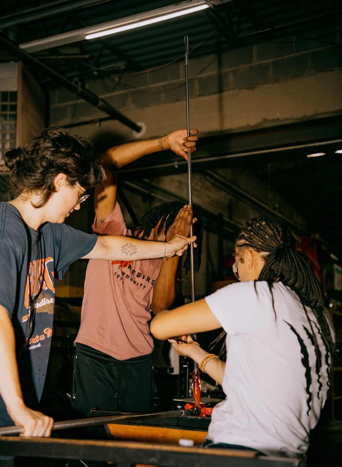 Three people working together to make glass art. One person is holding a long metal rod, and the other two are assisting with the process at a work table.