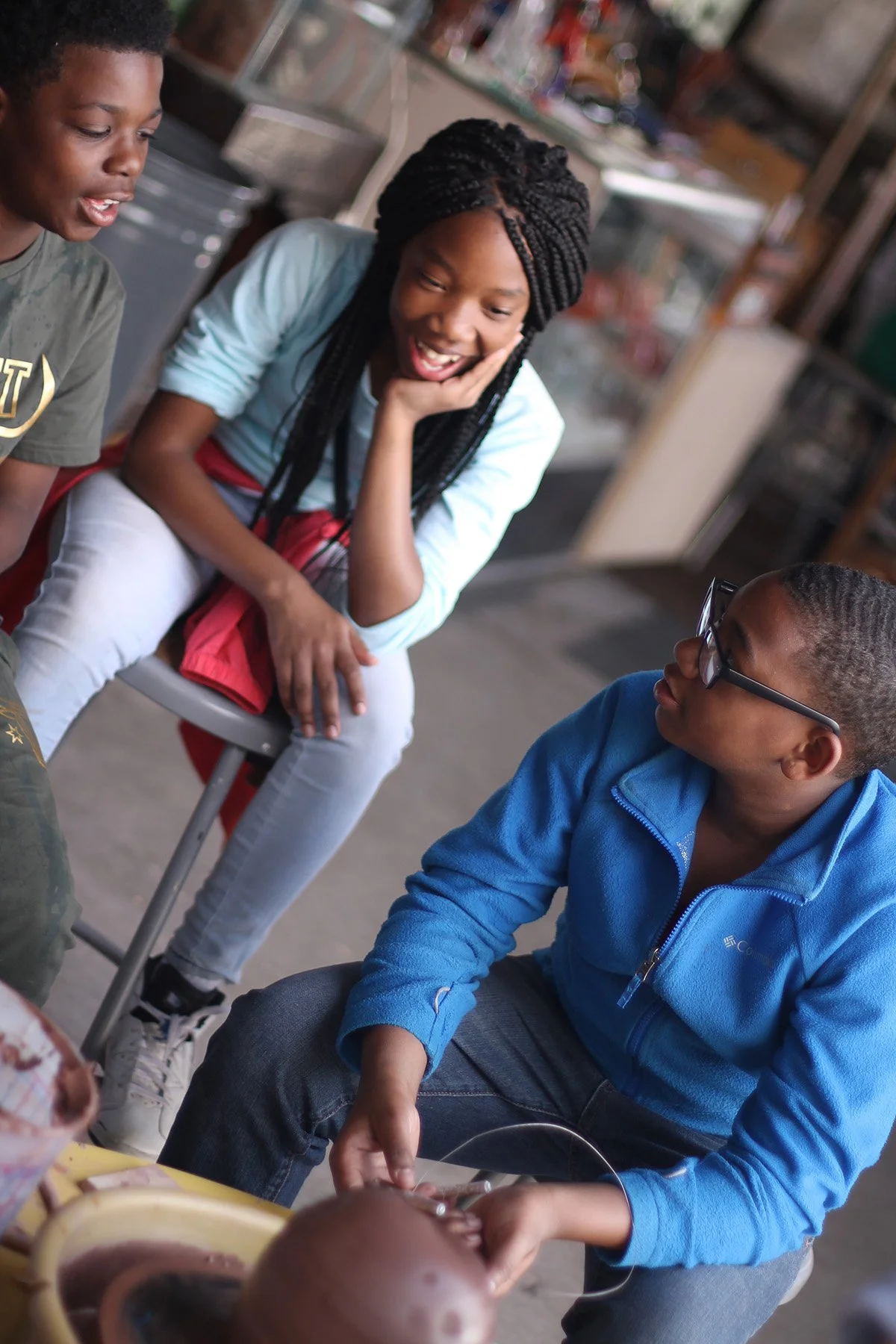 Three children, two boys and a girl, are gathered around a pottery wheel in a casual setting, engaging in conversation and sharing a joyful moment.