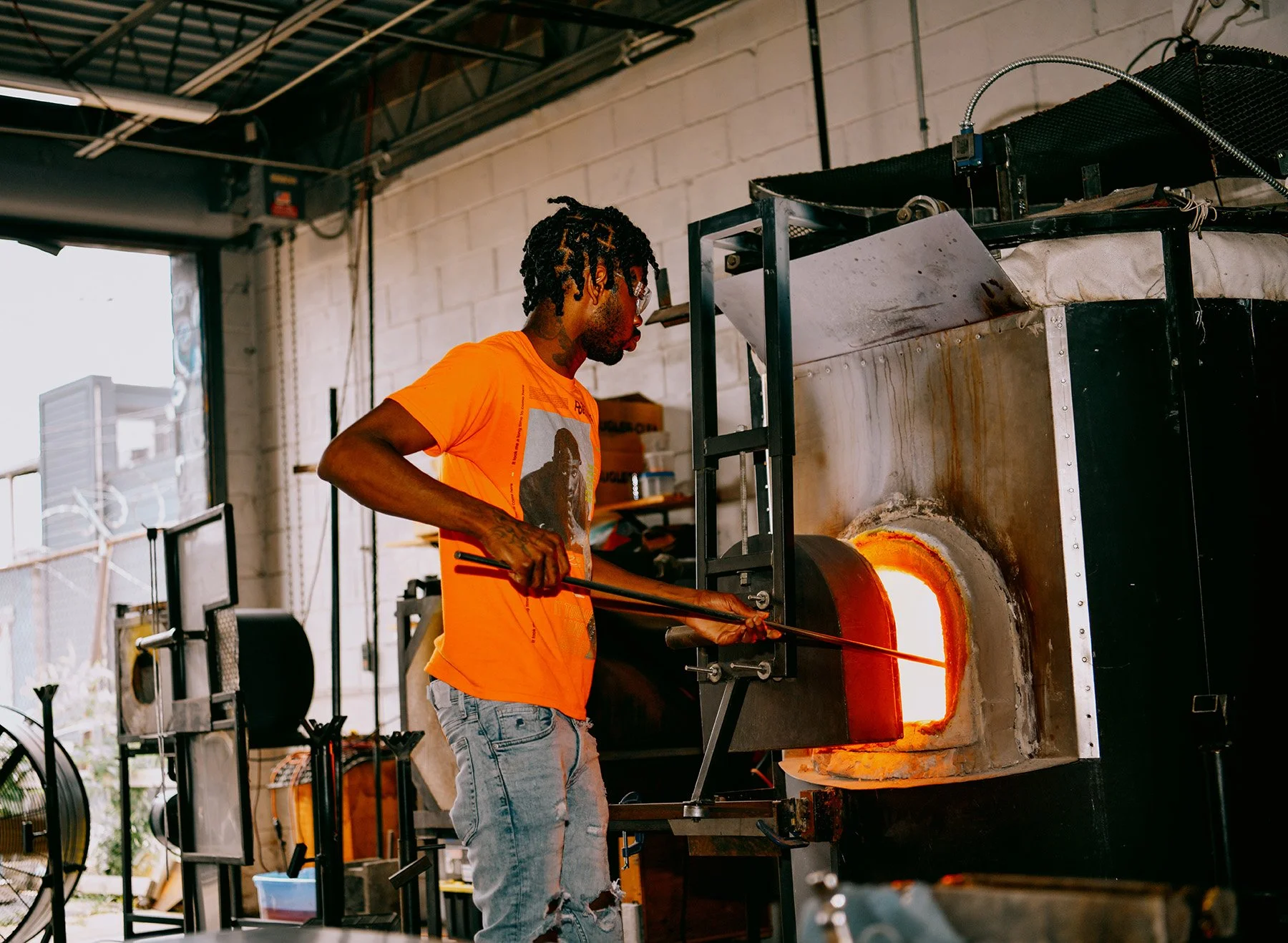 A man wearing an orange T-shirt with a graphic and ripped jeans is standing in front of a glass-blowing furnace, holding a long metal rod as he works with molten glass inside the furnace in a workshop.