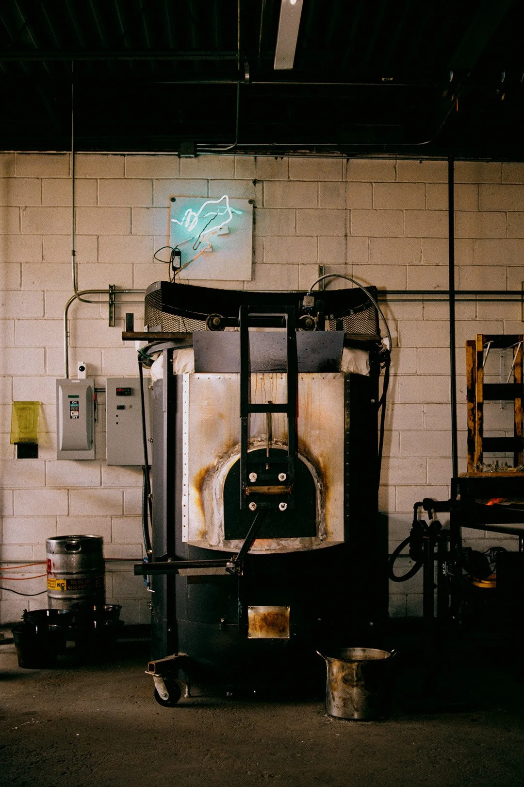 Glass kiln with a rusty front, sitting on a concrete floor in a studio with a brick wall. A neon sign of the Firebird logo is on the wall above.
