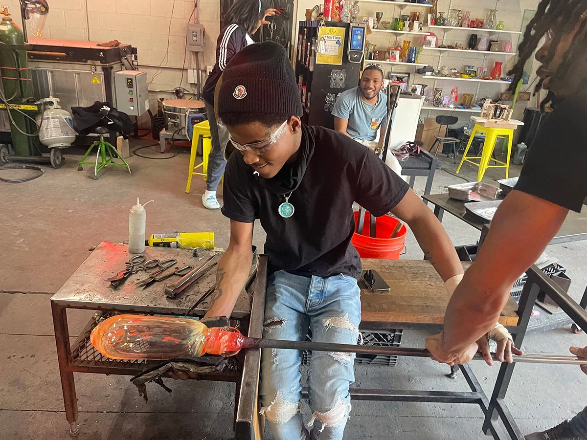 A person wearing glasses, a black beanie, and a black shirt is seated at a workbench with glassblowing tools, shaping molten glass into a shaped object, with other people and equipment visible in the background.