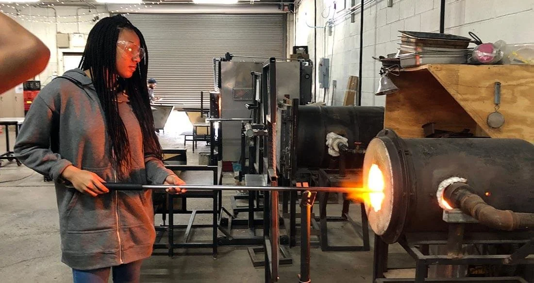 A woman in safety glasses and a hoodie watching glass blowing in a workshop with industrial equipment and a large furnace.