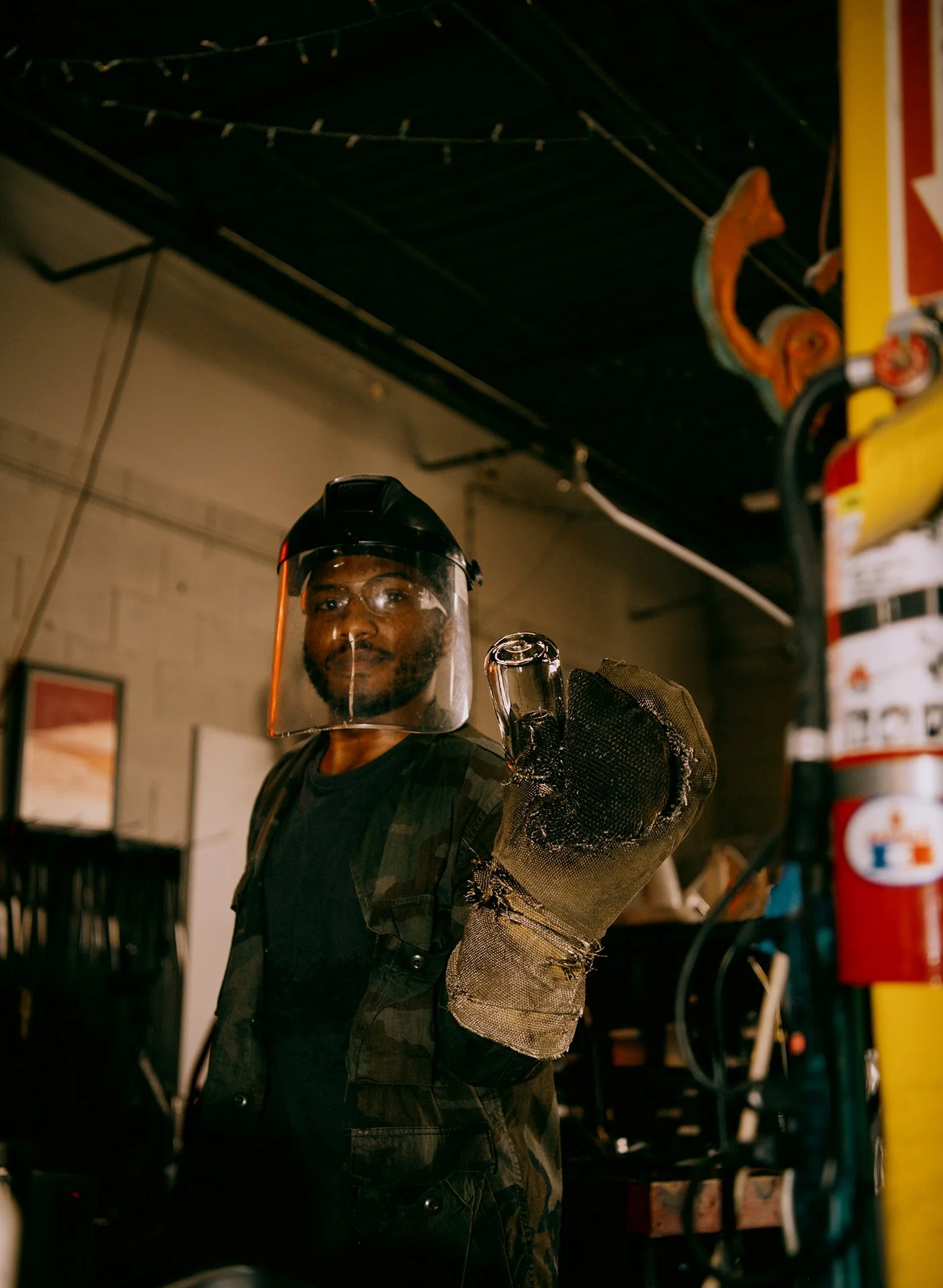 Man wearing a face shield and fireproof glove, giving a thumbs-up in a workshop or industrial setting.