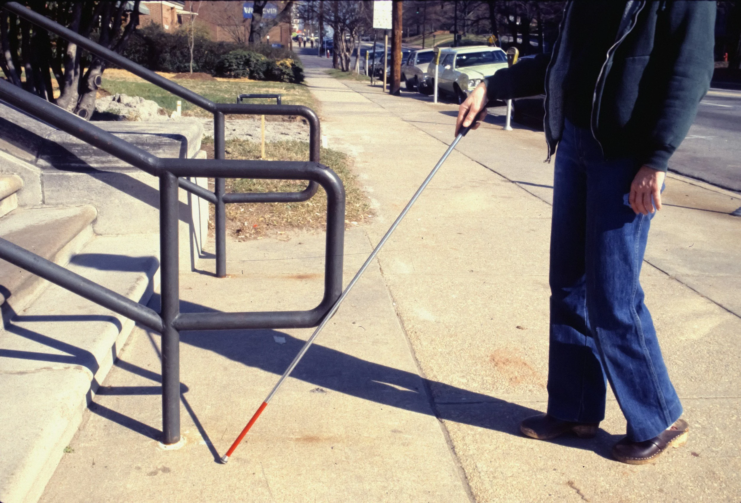 Person with a white cane using a cane at the sidewalk near stairs with handrails, preparing to navigate.