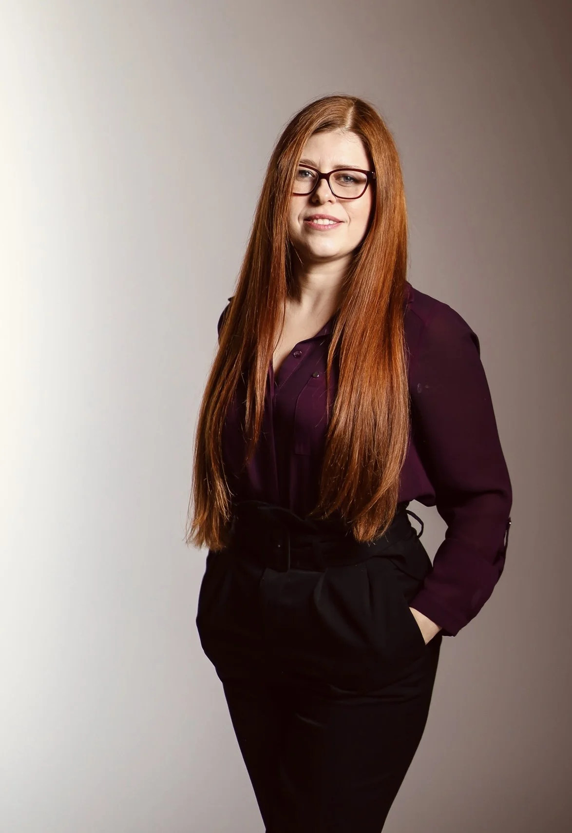 A woman with long red hair wearing glasses, a dark purple blouse, and black pants, standing with one hand in her pocket against a plain background.