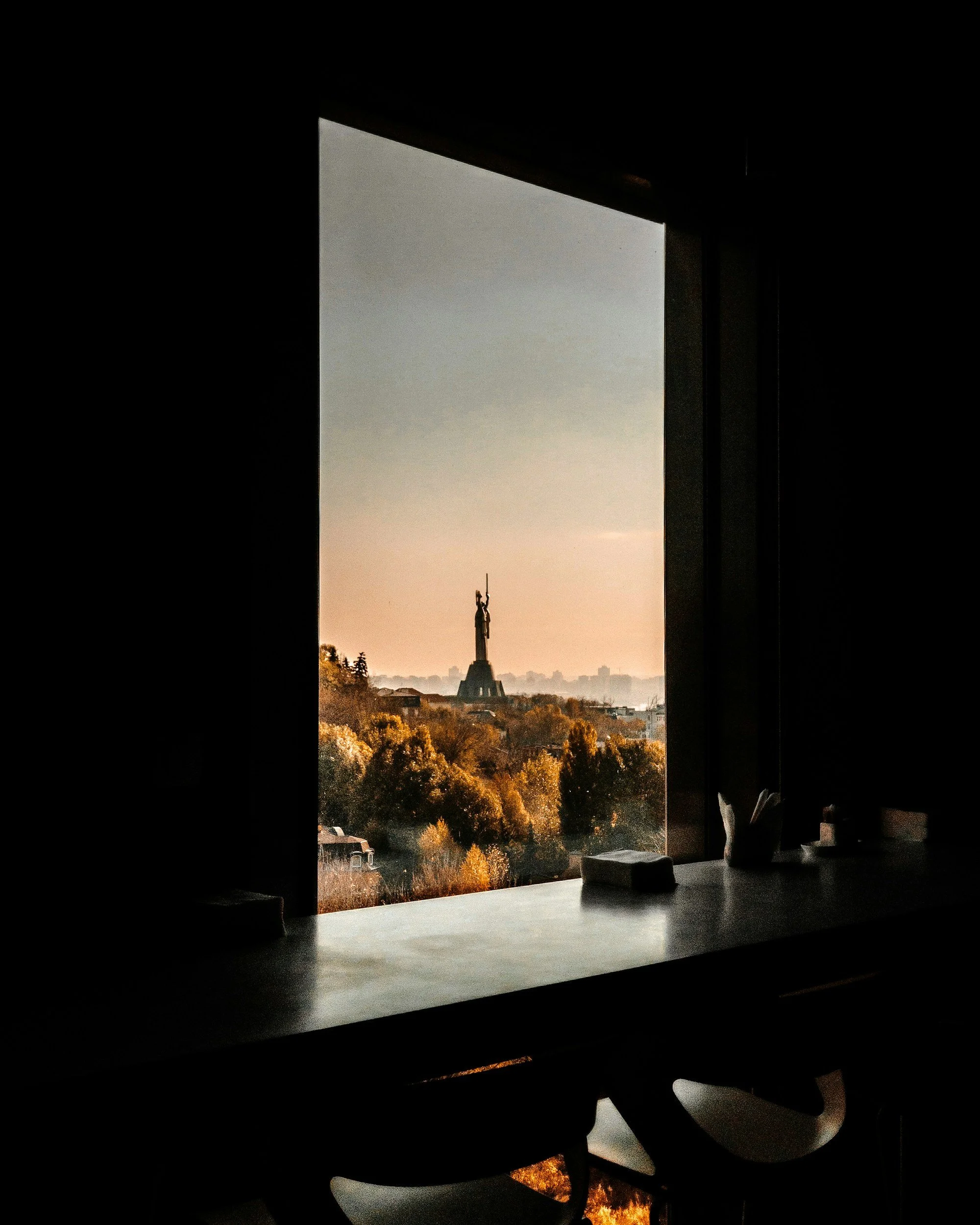 View of a landscape of Kyiv with trees and a monument with a statue in the distance, seen through a window from inside a dark room.