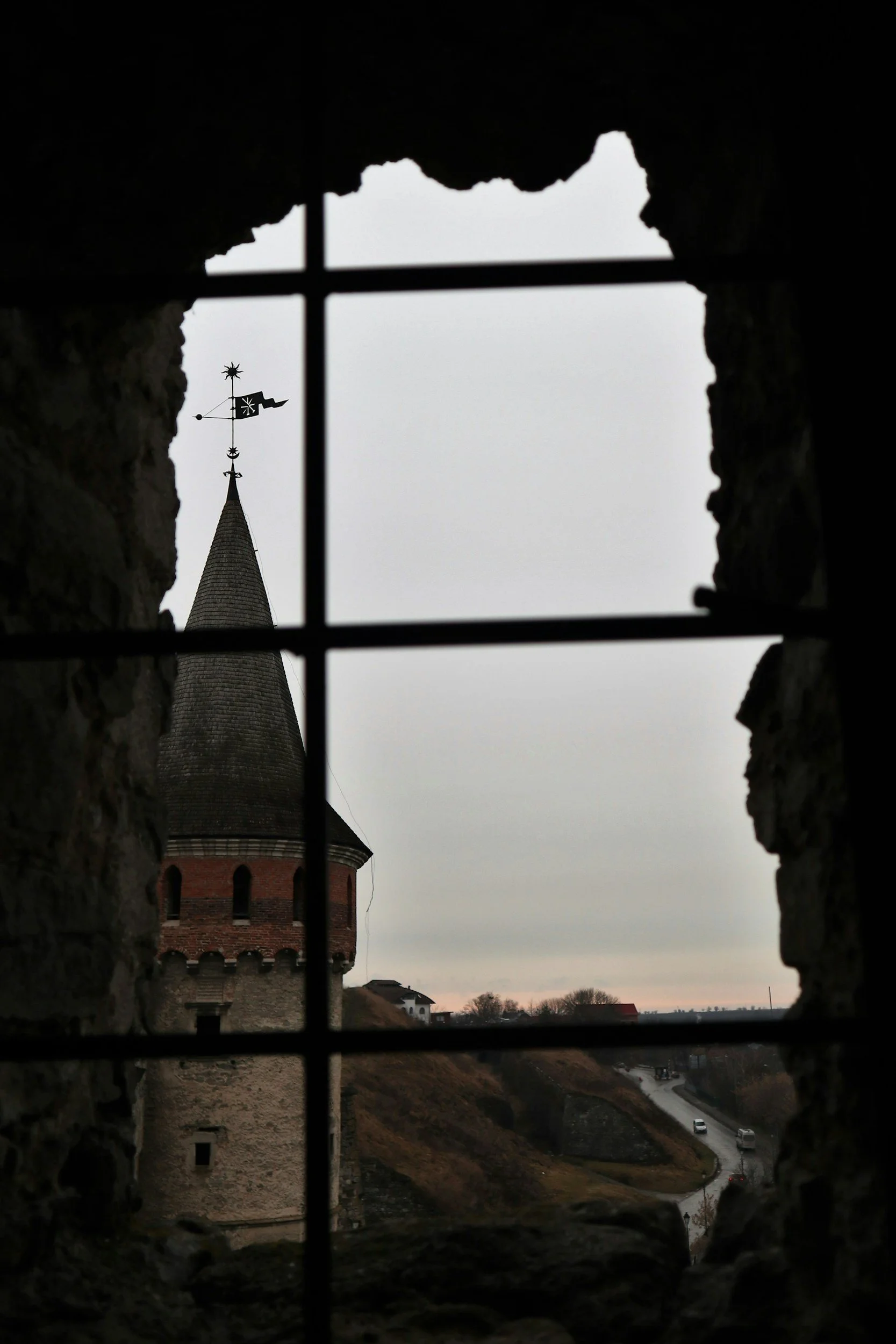 View of a church steeple with a weather vane through a small window with bars, in a cloudy sky.