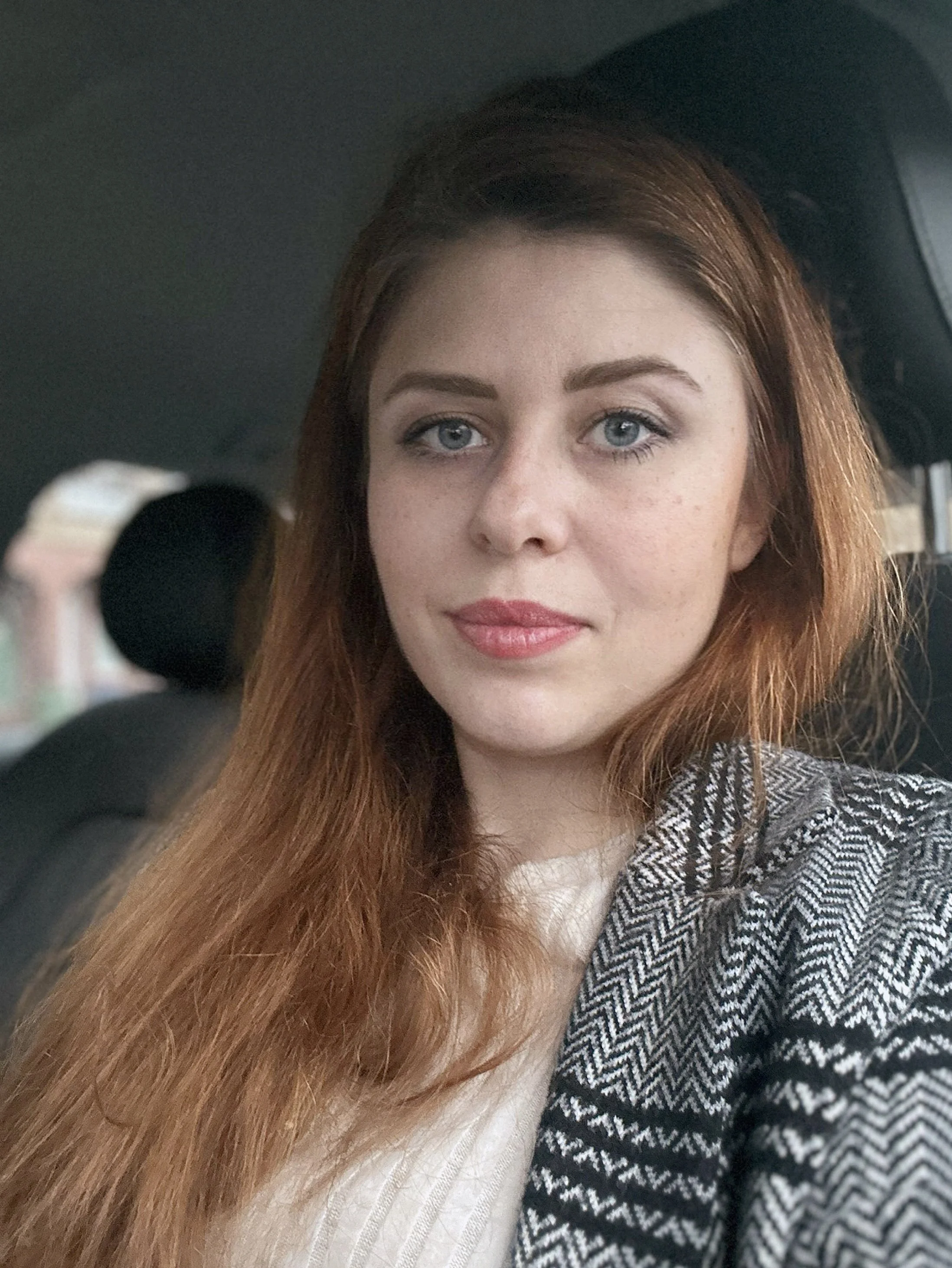 A woman with long red hair, blue eyes, and light makeup taking a selfie inside a car. She is wearing a herringbone patterned blazer over a light-colored top.