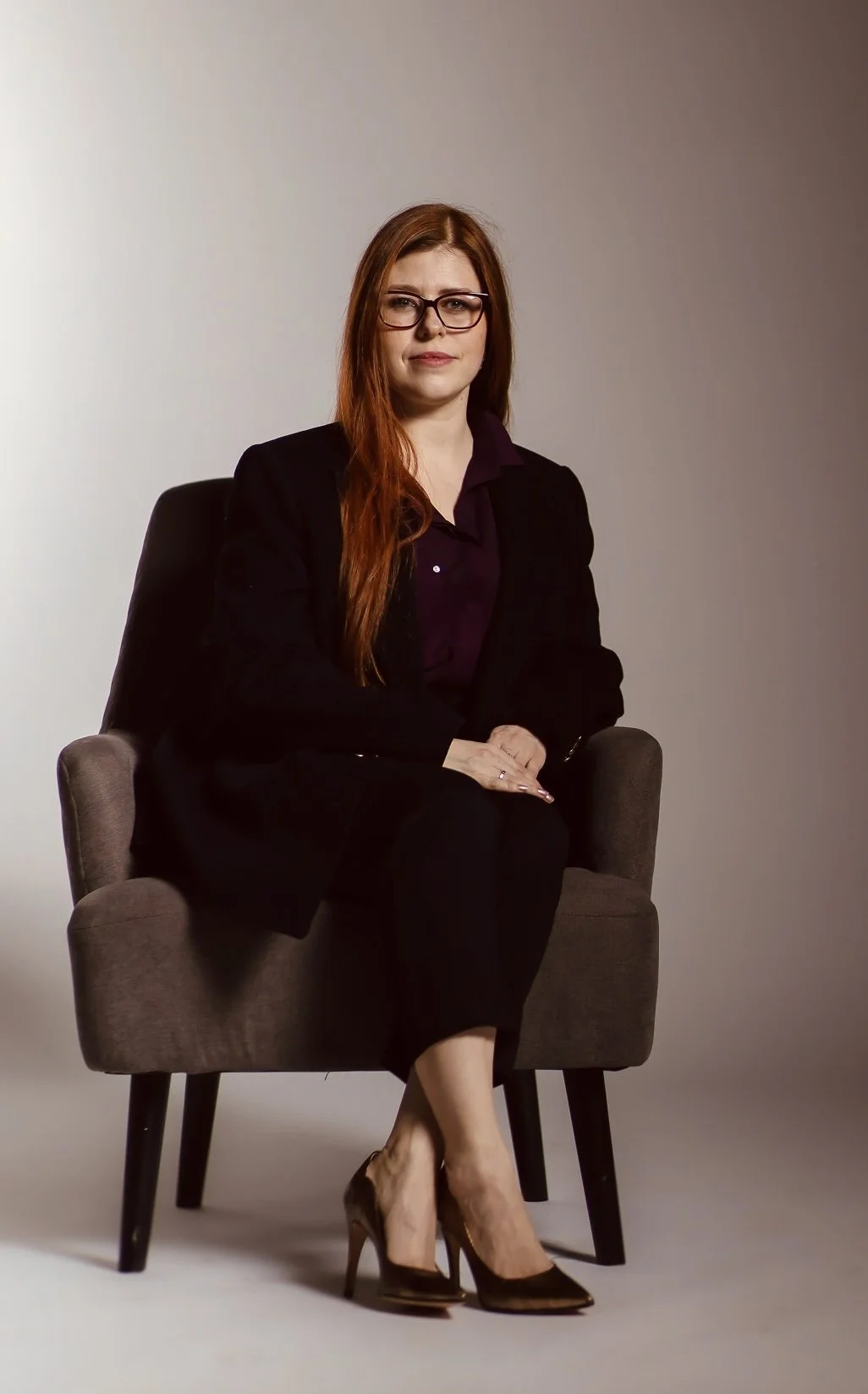 A woman with long red hair, wearing glasses, a black blazer, black pants, and black high heels, sitting on a gray chair against a plain gradient background.