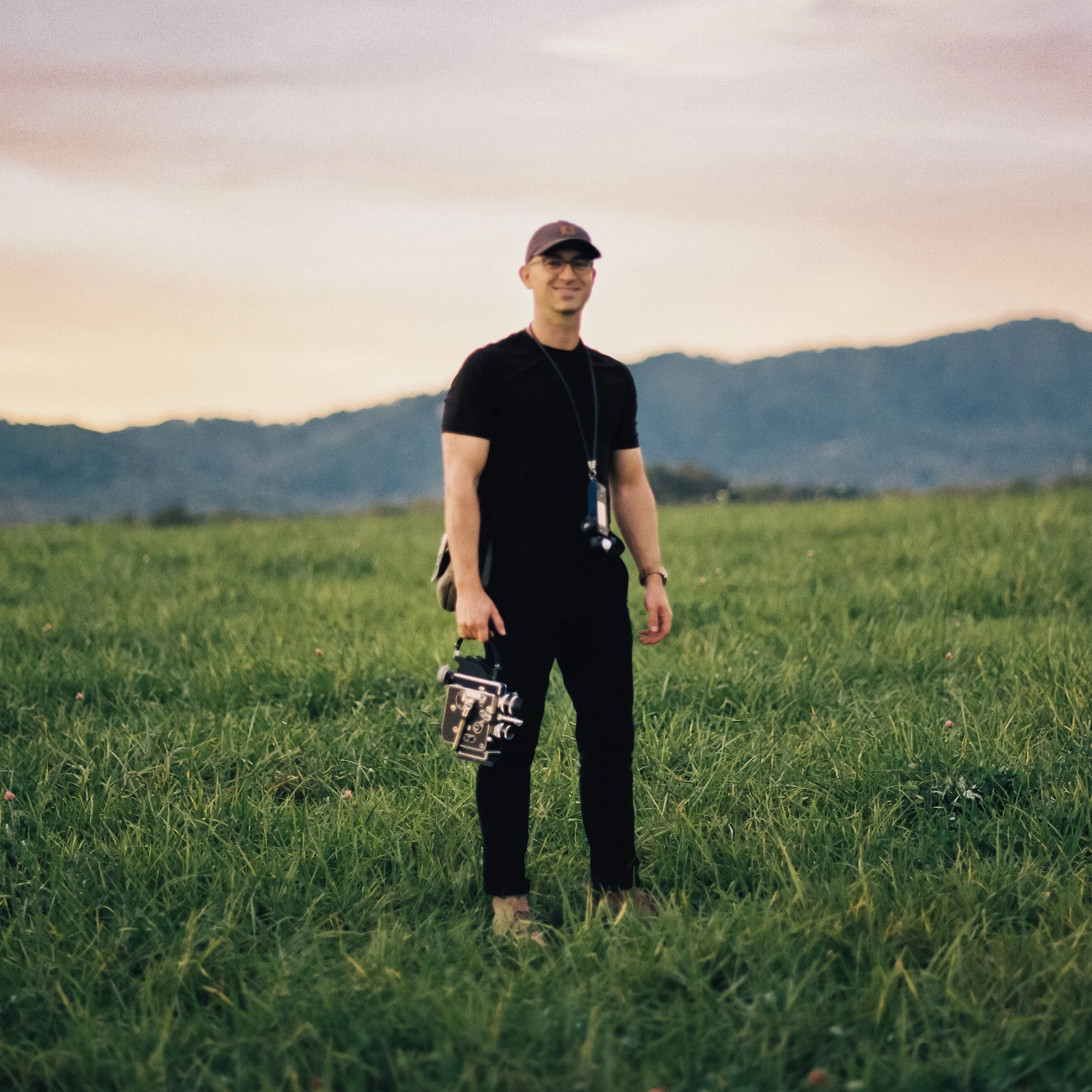 A man standing in a grassy field at sunset, holding a vintage camera, wearing glasses, a cap, and casual clothing.