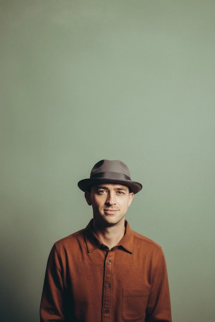 A man wearing a brown shirt and a fedora hat standing against a plain green background.