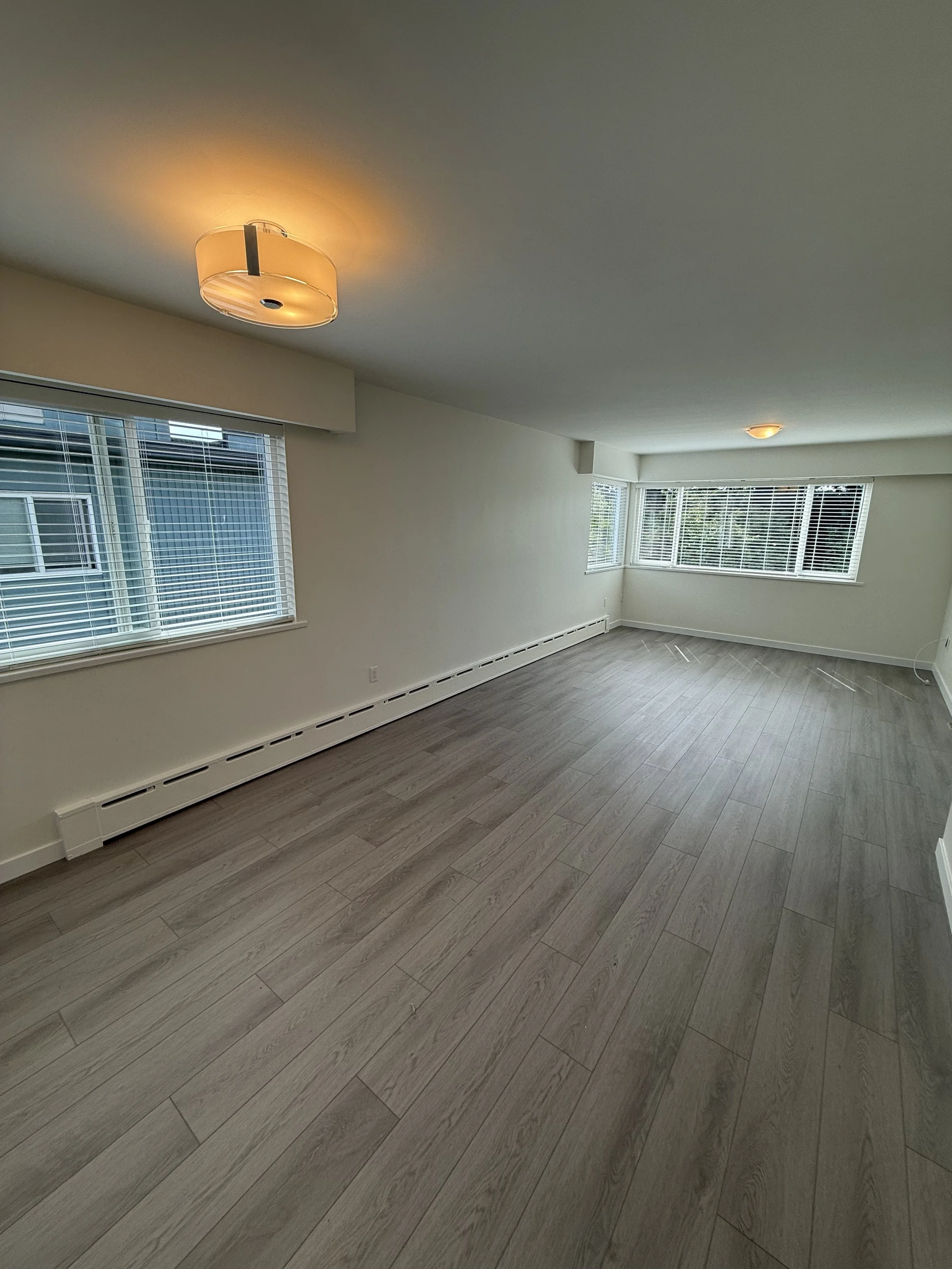 Empty living room with hardwood floors, white walls, multiple windows with blinds, and ceiling lights.