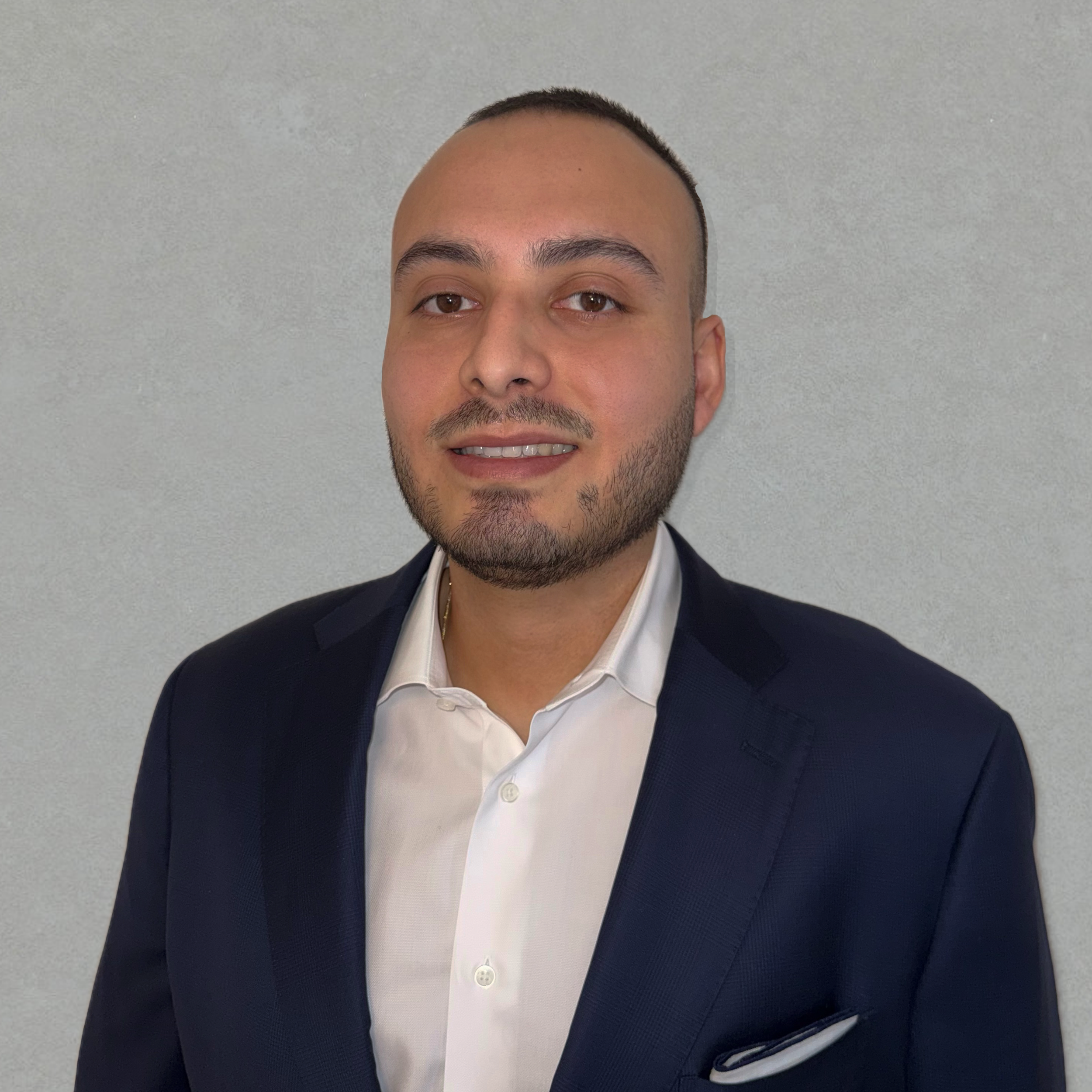 Headshot of a man wearing a navy suit jacket and a white shirt, standing against a plain grey background.