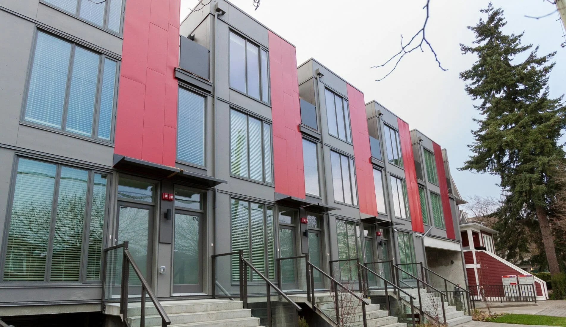 A modern multi-story apartment building with red and gray exterior, large glass windows, staircases leading to ground-level entrances, and a large tree nearby.