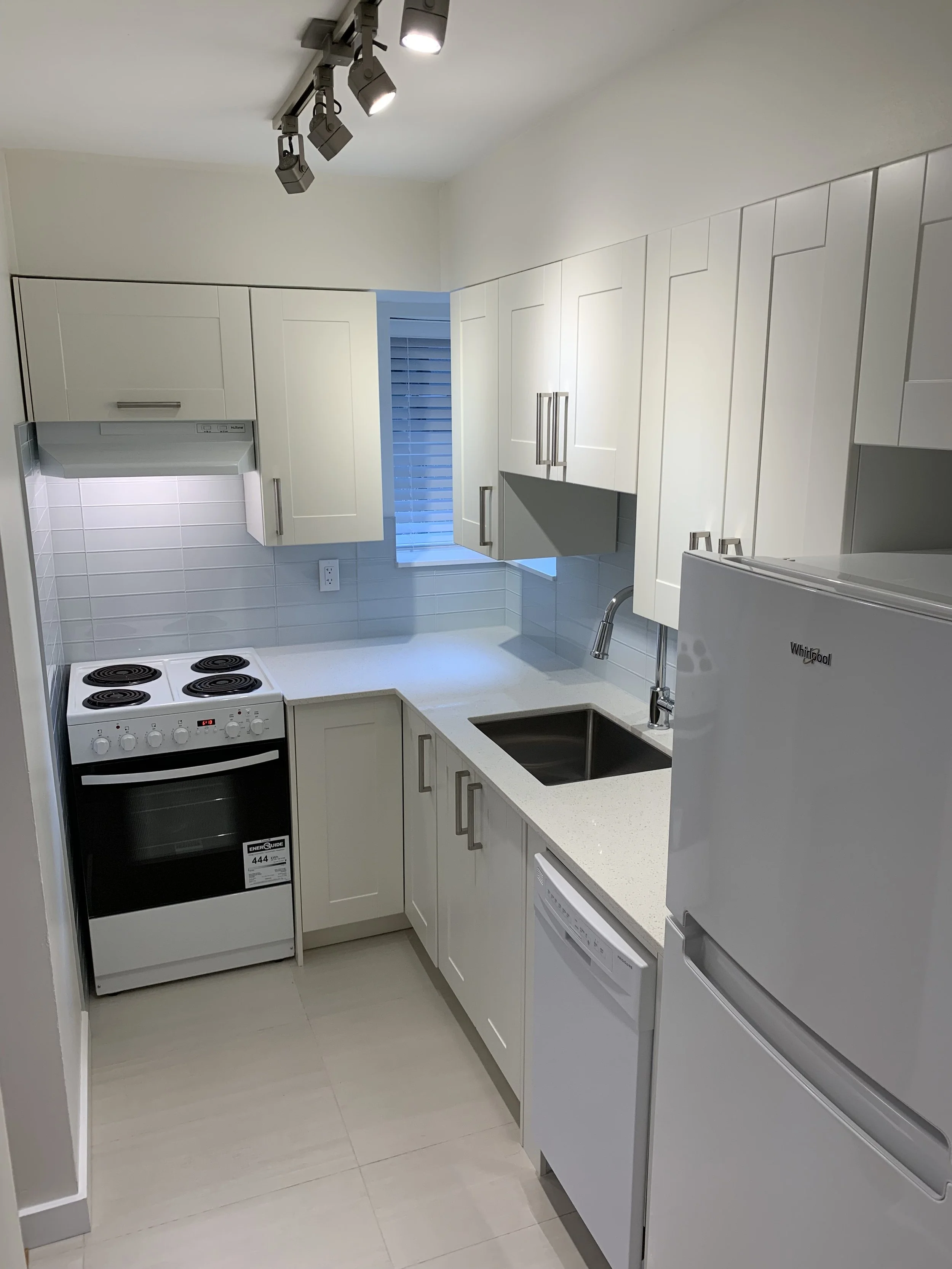 Small modern white kitchen with white cabinets, a white refrigerator, a white dishwasher, and a white stove, with a small window above the sink and track lighting on the ceiling.