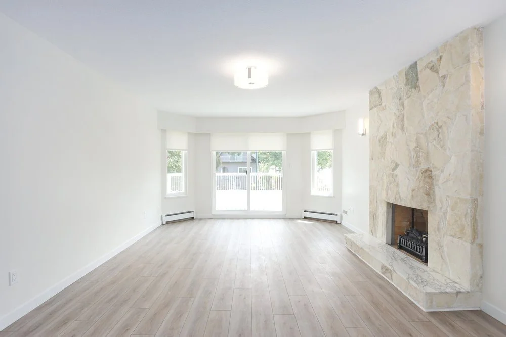 Empty living room with light-colored wood flooring, a large stone fireplace, and multiple windows letting in natural light.
