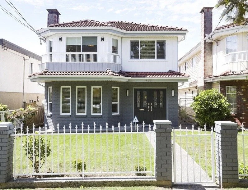 A two-story house with a dark blue lower level and white upper level, featuring a small front yard with a white fence, and a black front door flanked by windows.