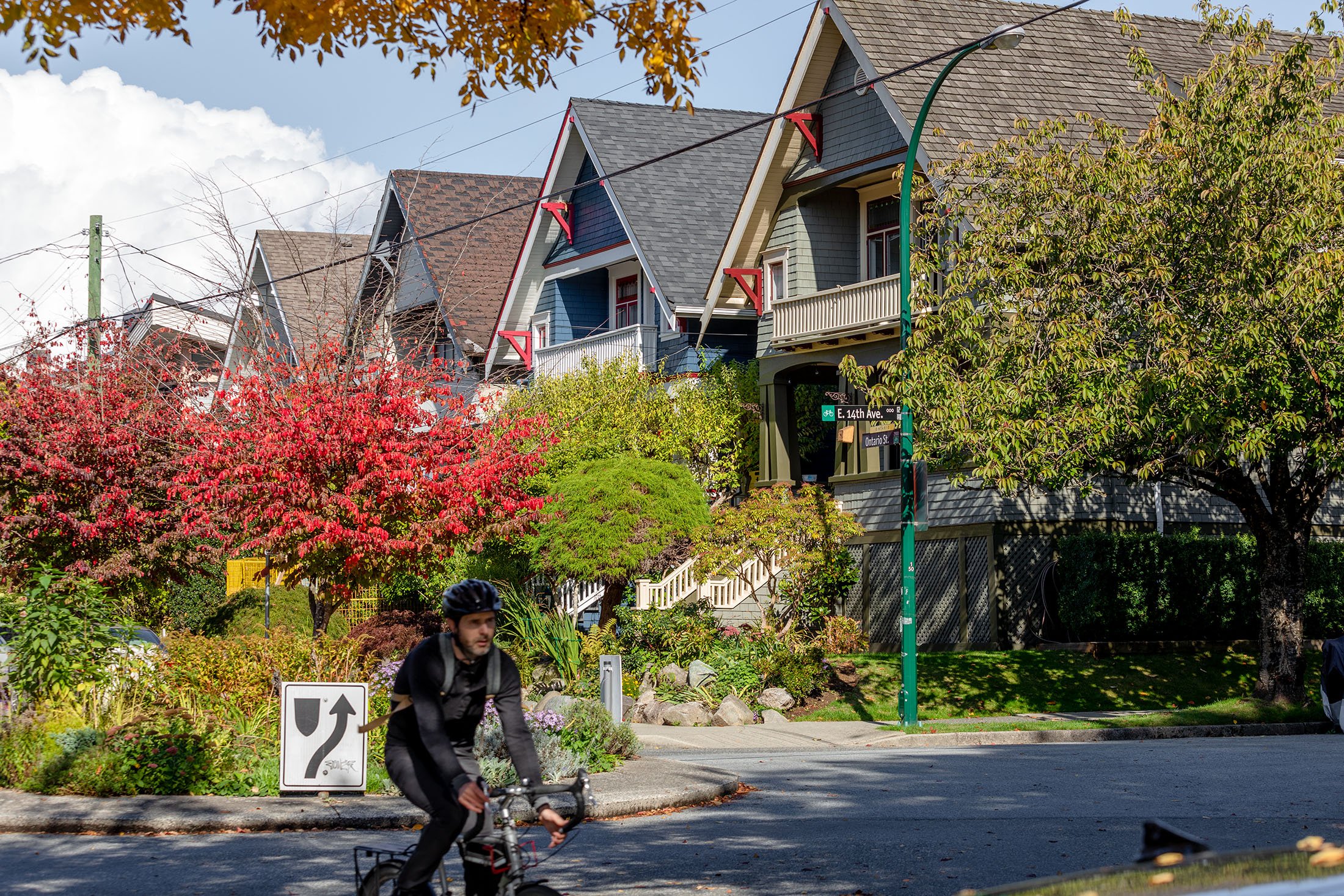 A man wearing a helmet riding a bicycle on a street with colorful houses and trees with autumn foliage; street signs indicate the intersection of E. 14th Avenue and Ontario Street.