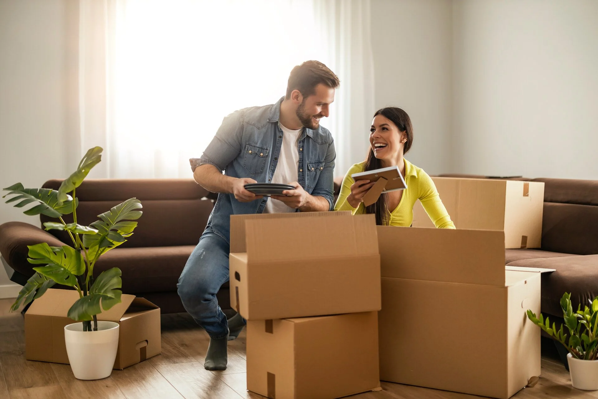 A smiling couple moving into a new home, surrounded by unpacked boxes and houseplants in a bright living room.
