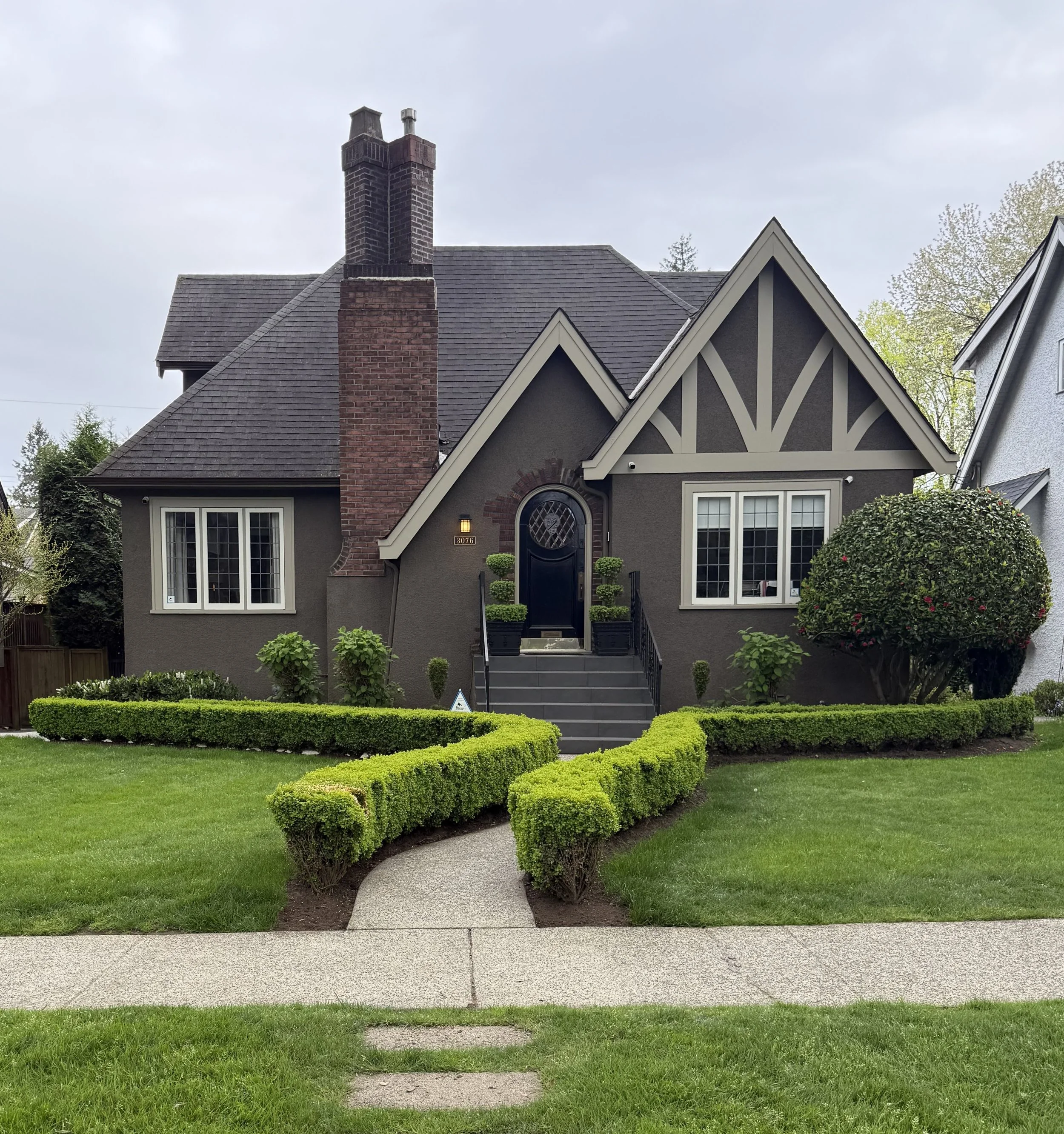 Front view of a dark gray house with a steep roof, brick chimney, white-trimmed windows, and a black front door. The house has a well-manicured lawn, a concrete walkway, and neatly trimmed bushes and trees.