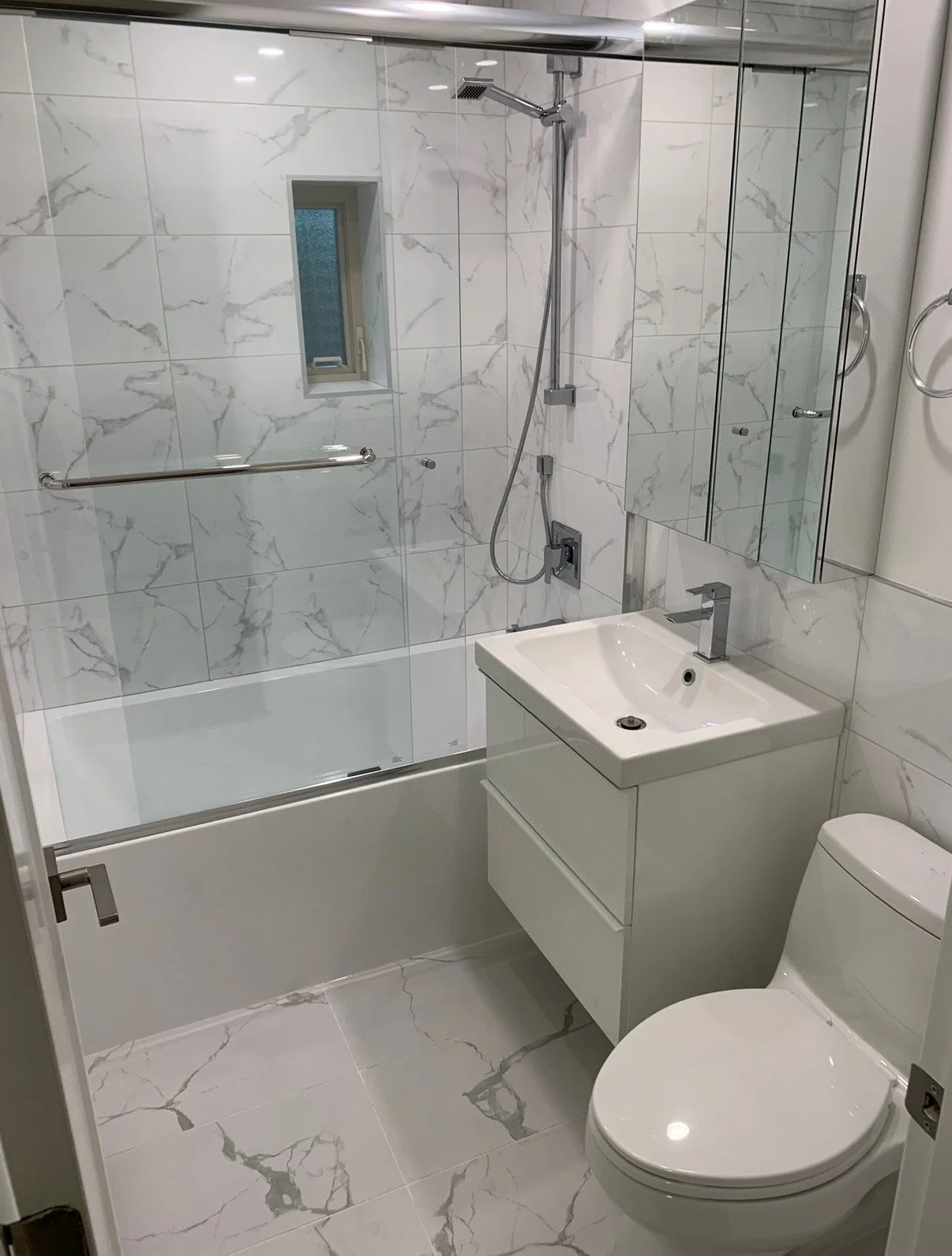A modern bathroom with a white marble tile design, a glass-enclosed shower, a small window, a white wall-mounted sink with a silver faucet, a mirror cabinet, and a white toilet.