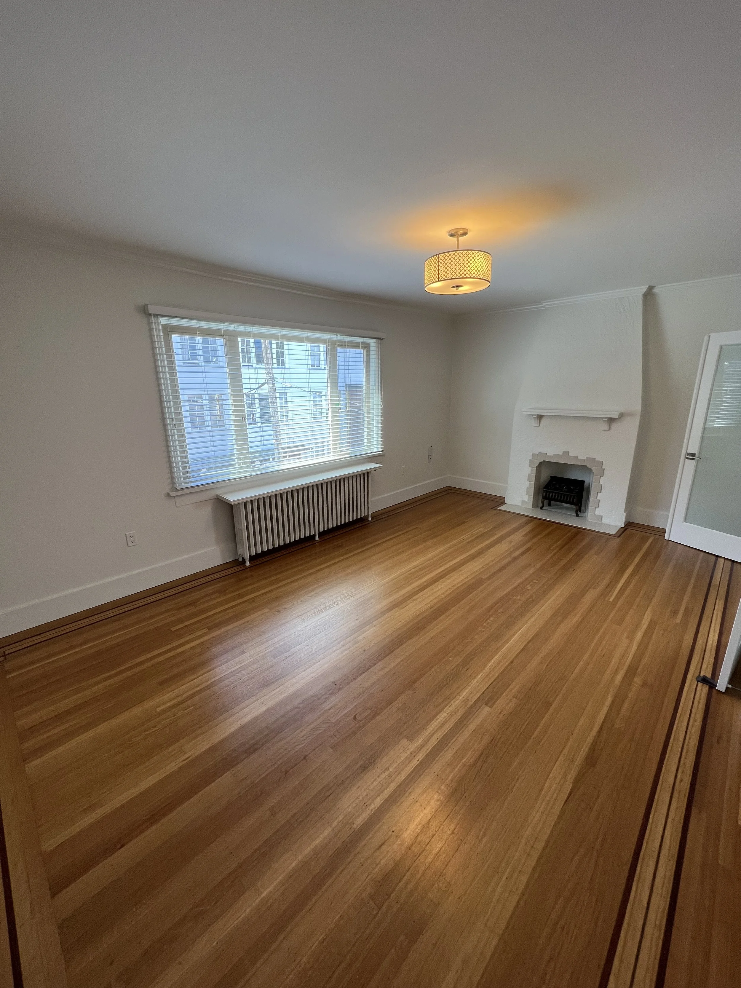 Empty living room with hardwood floors, white walls, a large window with blinds, a white fireplace, and a ceiling light fixture.