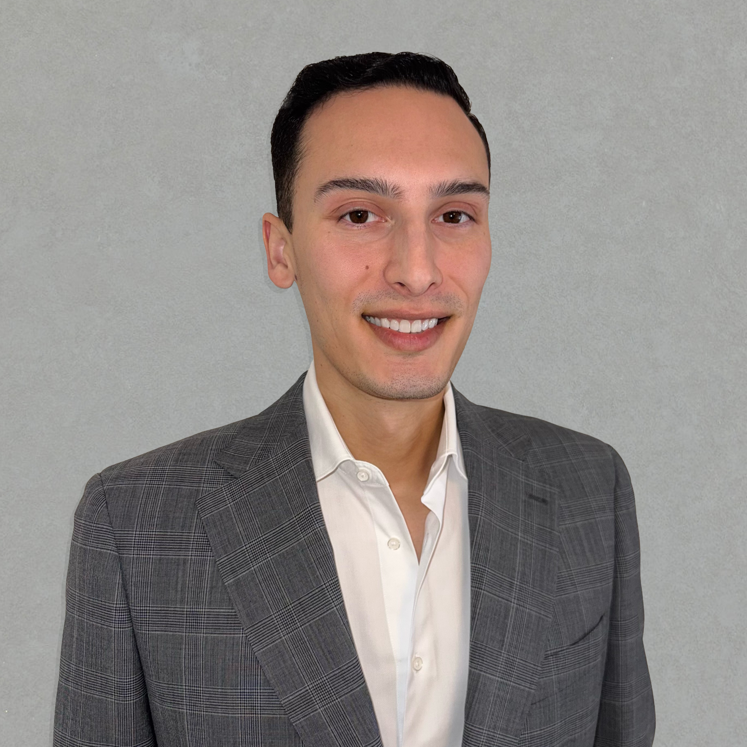 A young man in a checked gray blazer and white shirt smiling at the camera against a plain gray background.