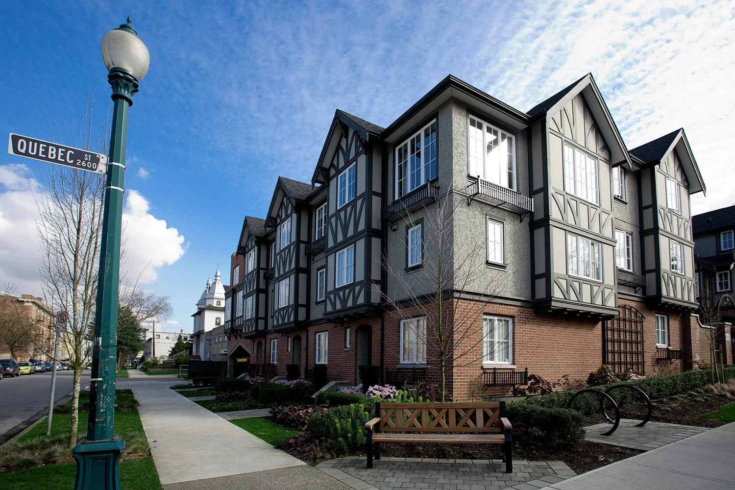 A modern multi-story residential building with gray and brick exterior, large windows, and small balconies, located at the corner of Quebec Street in a neighborhood with a sidewalk, trees, and a park bench under a partly cloudy sky.