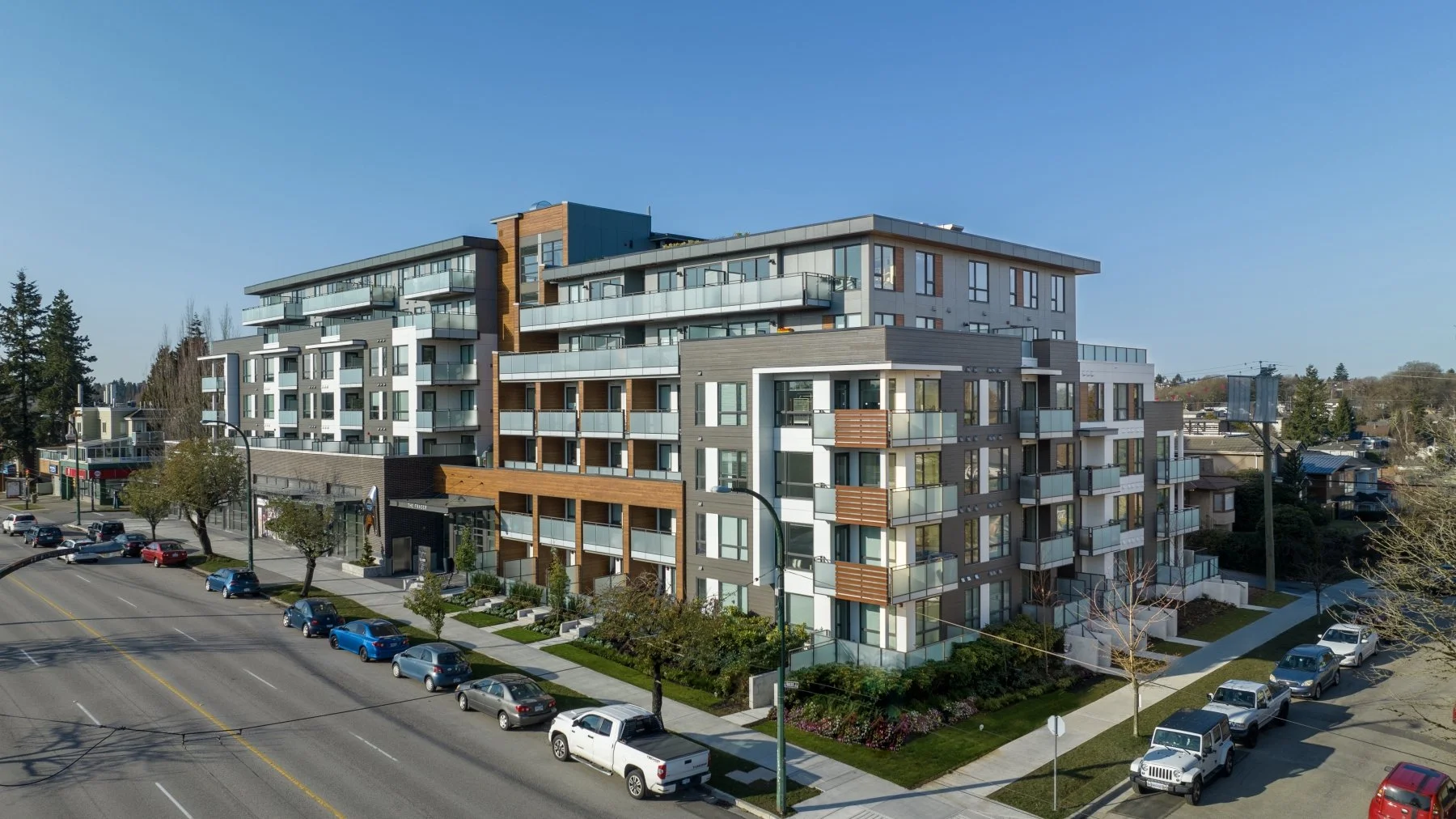 Modern multi-story apartment building with glass balconies and landscaped greenery, situated beside a busy street with parked cars.