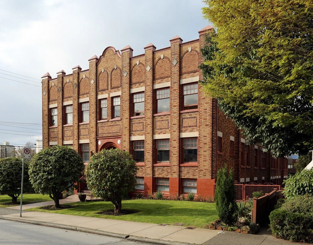 A three-story brick building with decorative architectural details, large rectangular windows, and a landscaped front yard with trimmed bushes and a sidewalk in an urban setting.