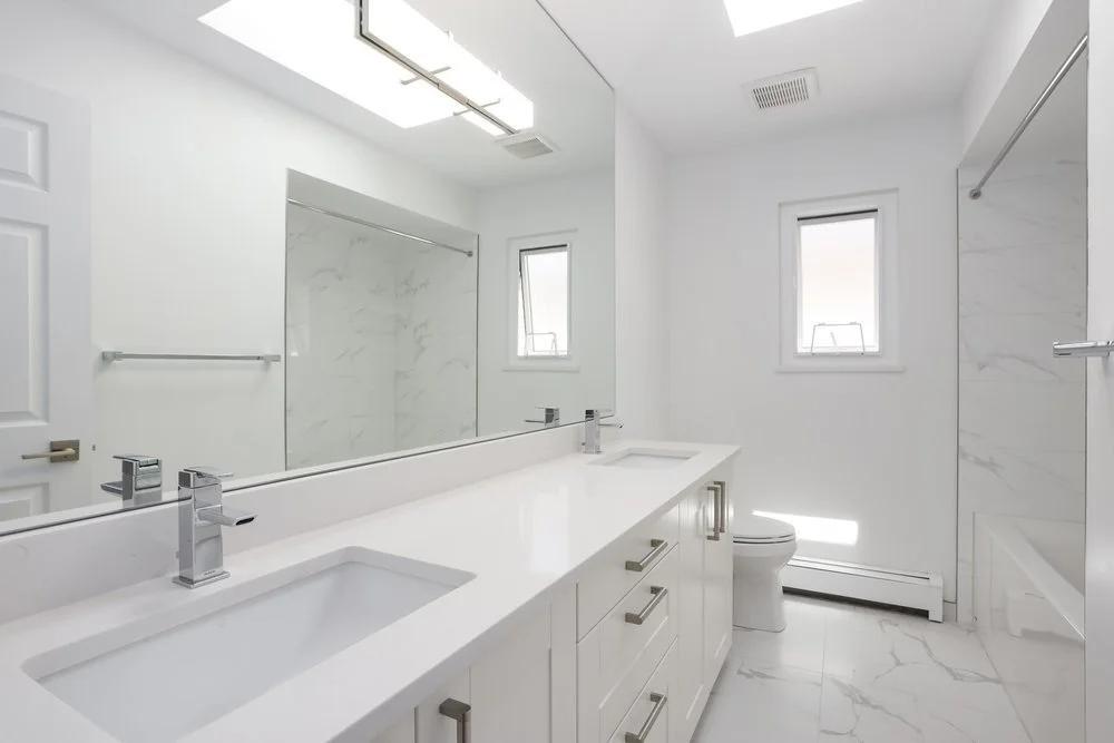 A modern, white bathroom with a double sink vanity, large mirror, two small windows, marble flooring, and a shower area with a glass door.