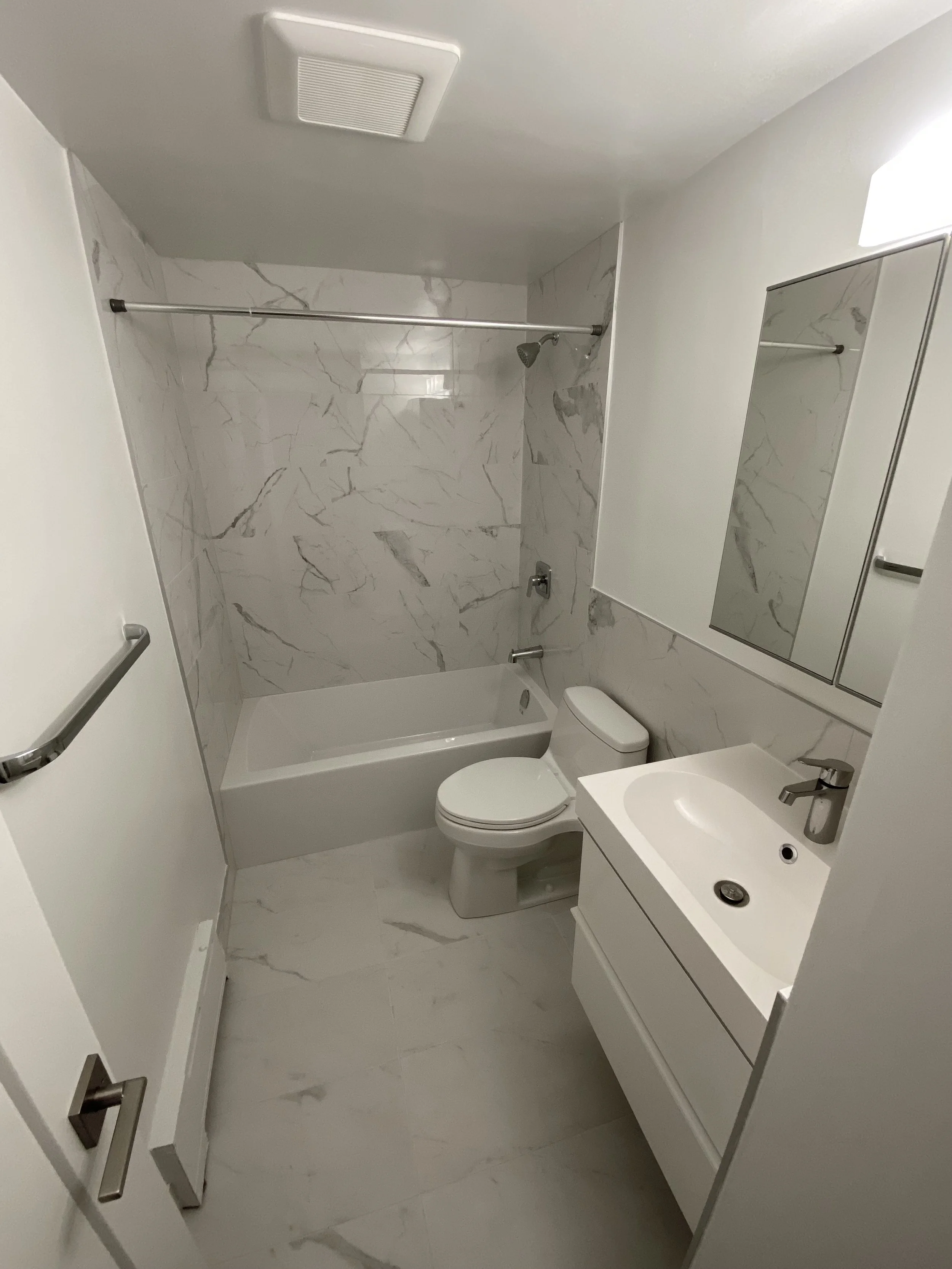 Small bathroom with white marble walls, a bathtub with shower, toilet, and a tiny white vanity with a sink and mirror.