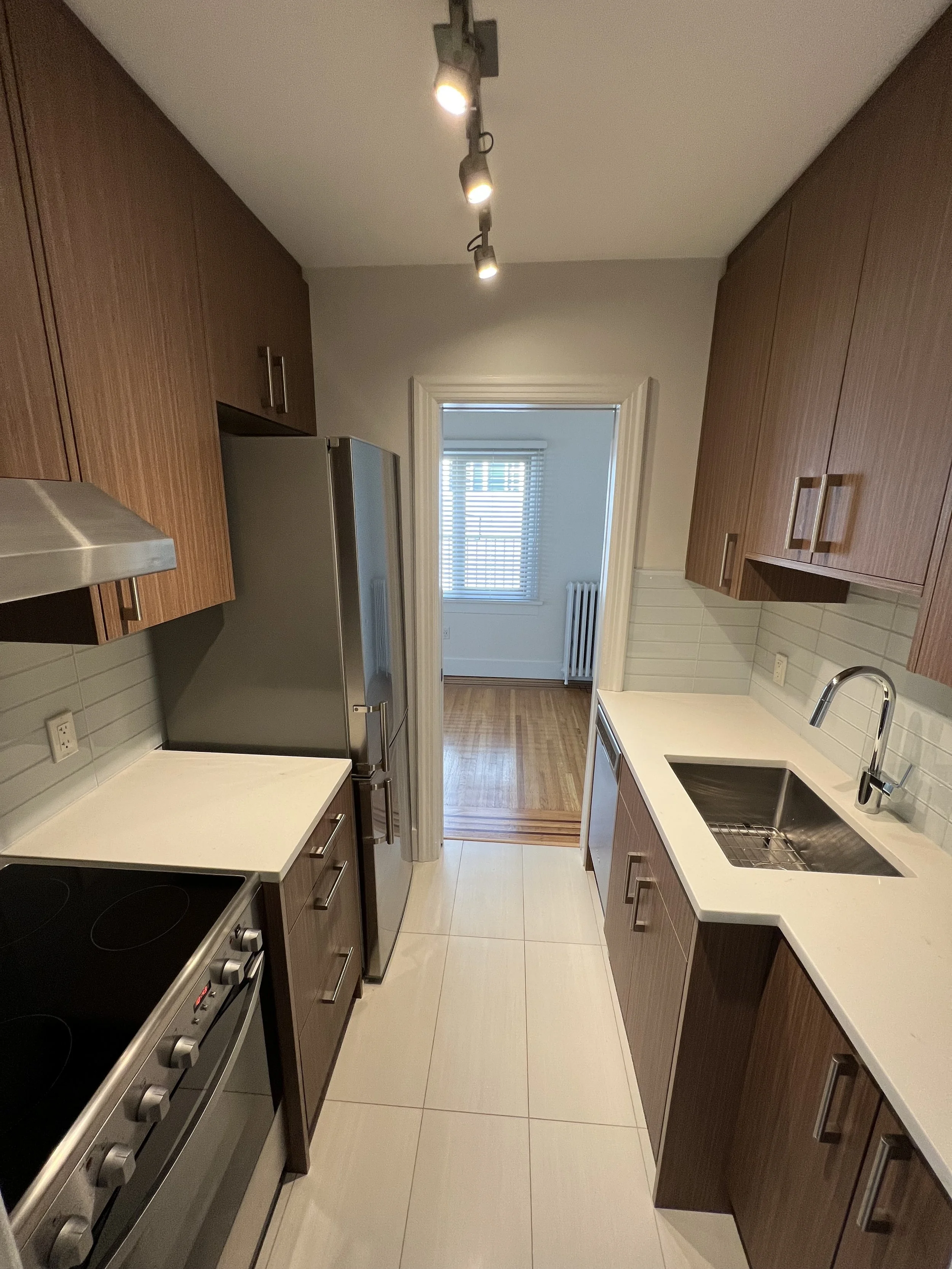 Modern narrow galley kitchen with wooden cabinetry, white countertops, stainless steel appliances, and a view into a bright room with wooden floors and window blinds.