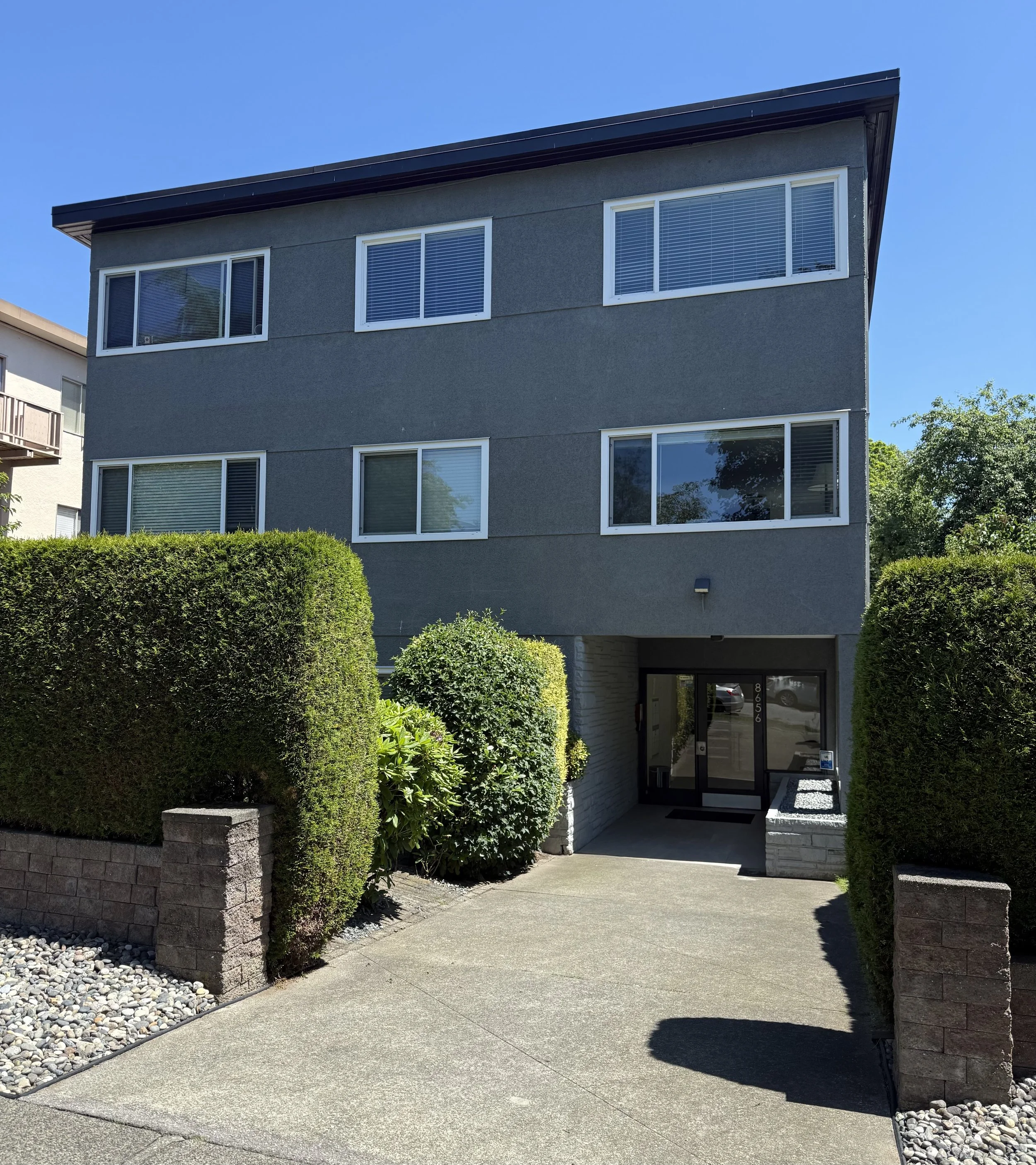 Three-story gray apartment building with white-framed windows and a driveway entrance, surrounded by green bushes and trees under a clear blue sky.