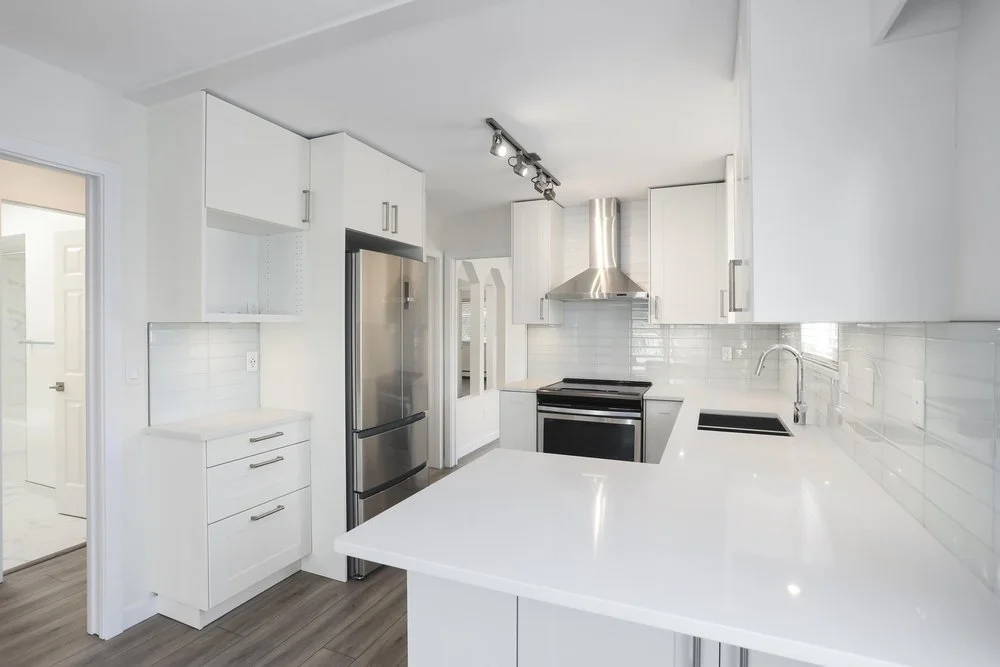Modern white kitchen with stainless steel refrigerator, oven, and range hood, white cabinets, and a white countertop.